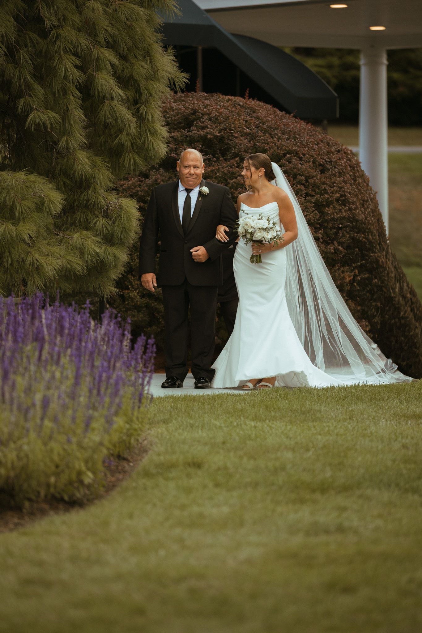 Bride in white gown and veil walks with her father through a garden toward the wedding ceremony.