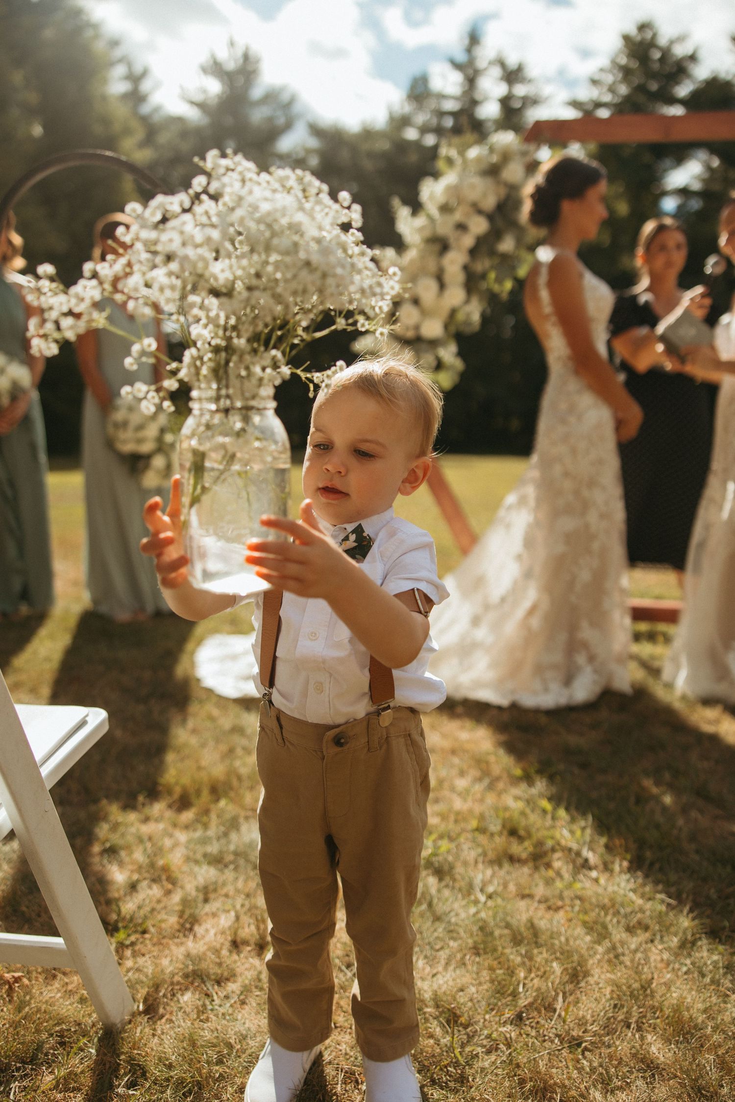 Adorable ring bearer in suspenders holds glass jar of baby's breath flowers at outdoor wedding ceremony.