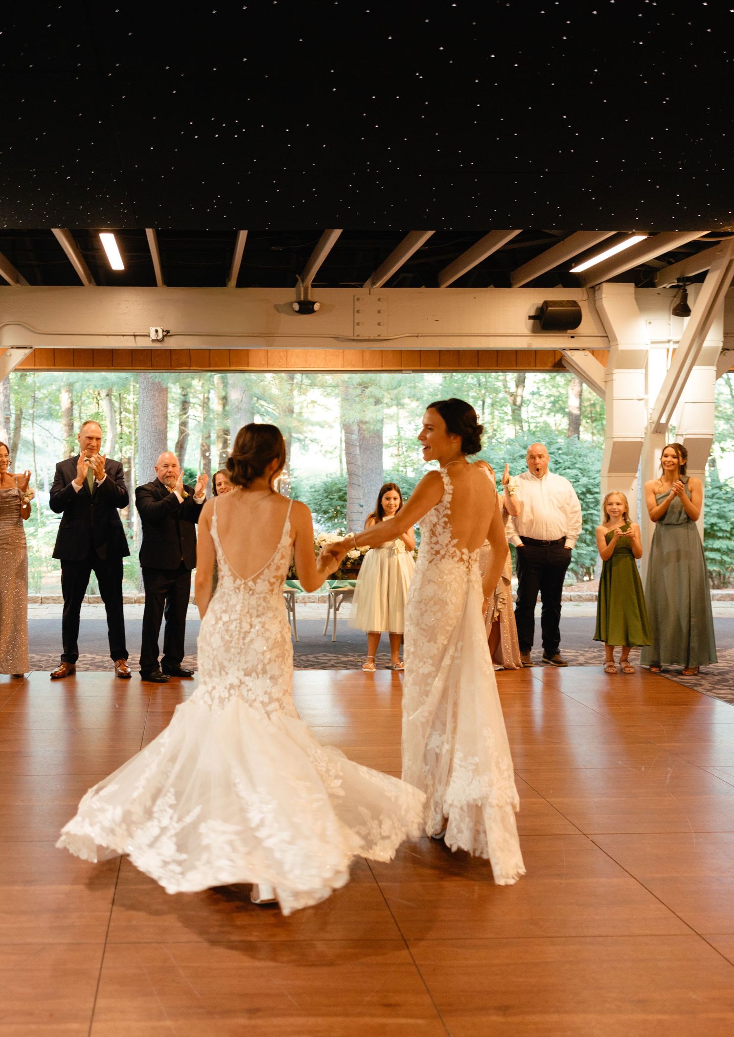 Two brides in lace wedding gowns sharing first dance while guests applaud at indoor reception.film-inspired