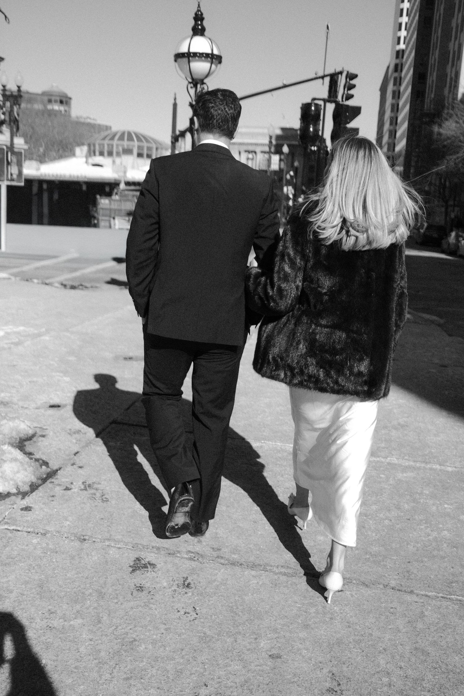 Black and white photo of a well-dressed couple walking away down a sunny city sidewalk casting long shadows.film-inspired