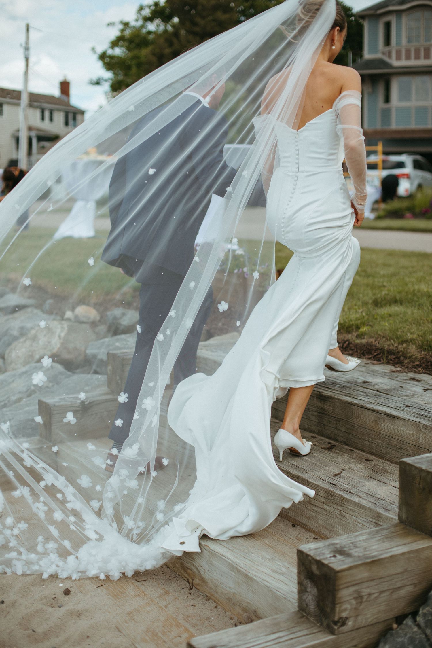 Bride in a fitted white gown with a floral cathedral veil climbs wooden steps with groom at coastal wedding.