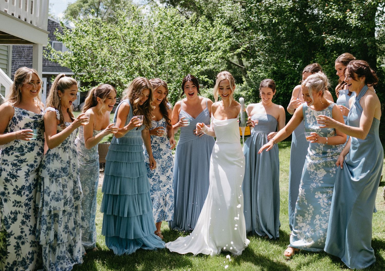 Bride in white gown celebrating with bridesmaids in mixed blue floral dresses outdoors on sunny day.