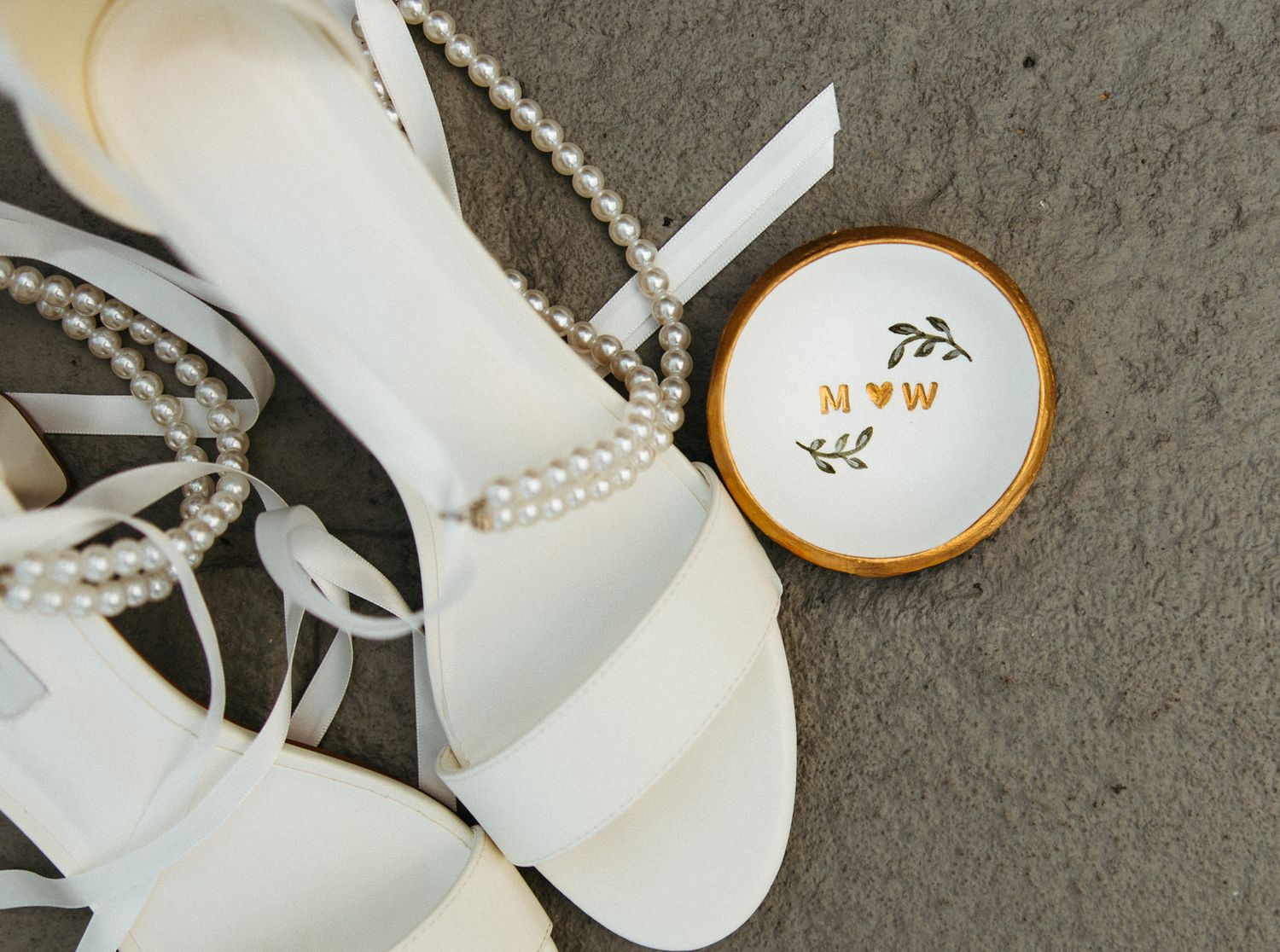 Close-up of white pearl-strap bridal heels beside personalized monogram ring dish on stone.