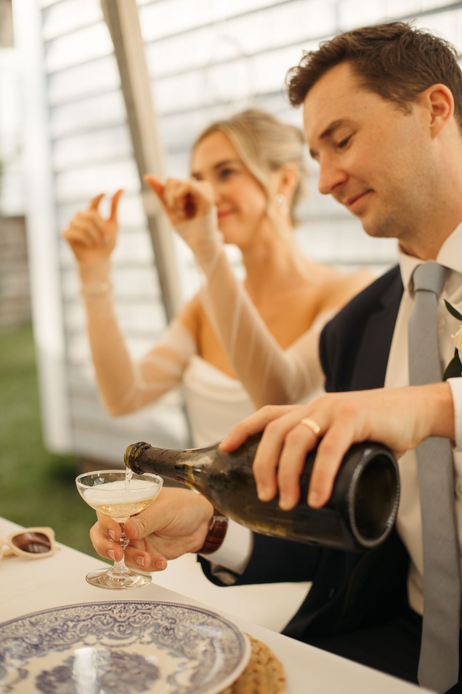 Groom pouring champagne into coupe glass beside smiling bride at outdoor wedding reception.