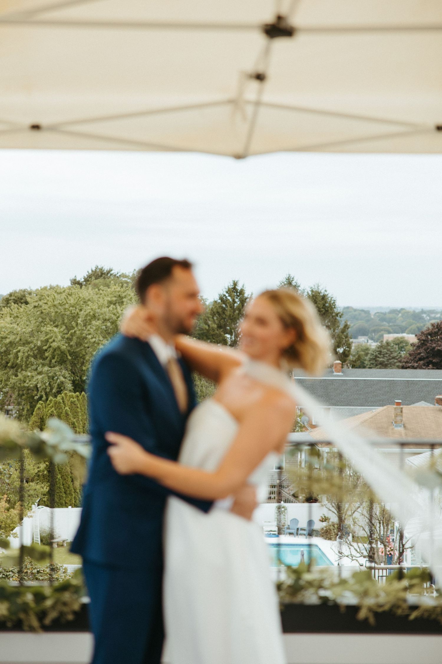 Bride and groom share romantic embrace on rooftop terrace with garden and pool views behind them.