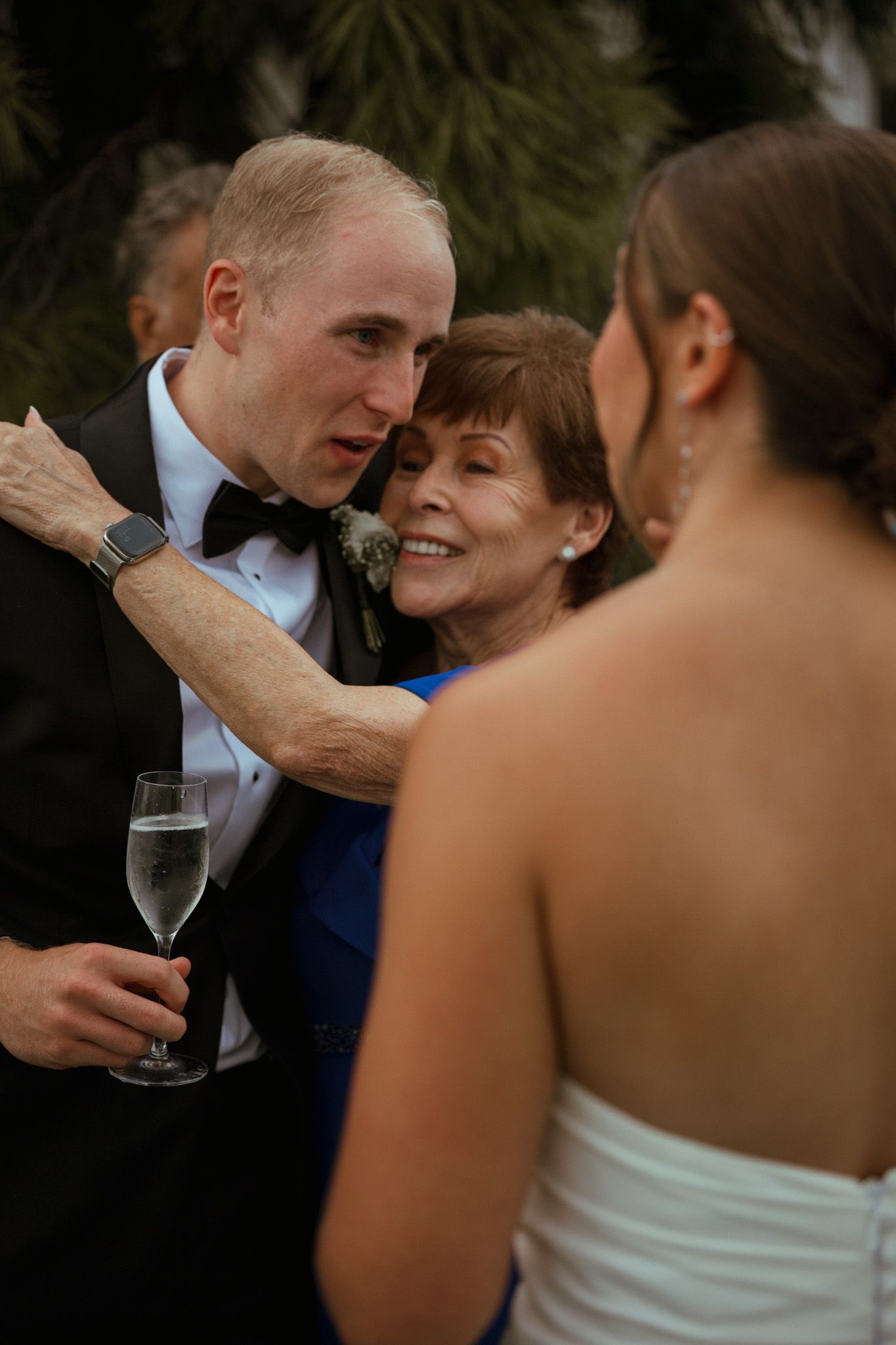 Groom embraces smiling older woman while holding champagne glass at outdoor wedding reception.