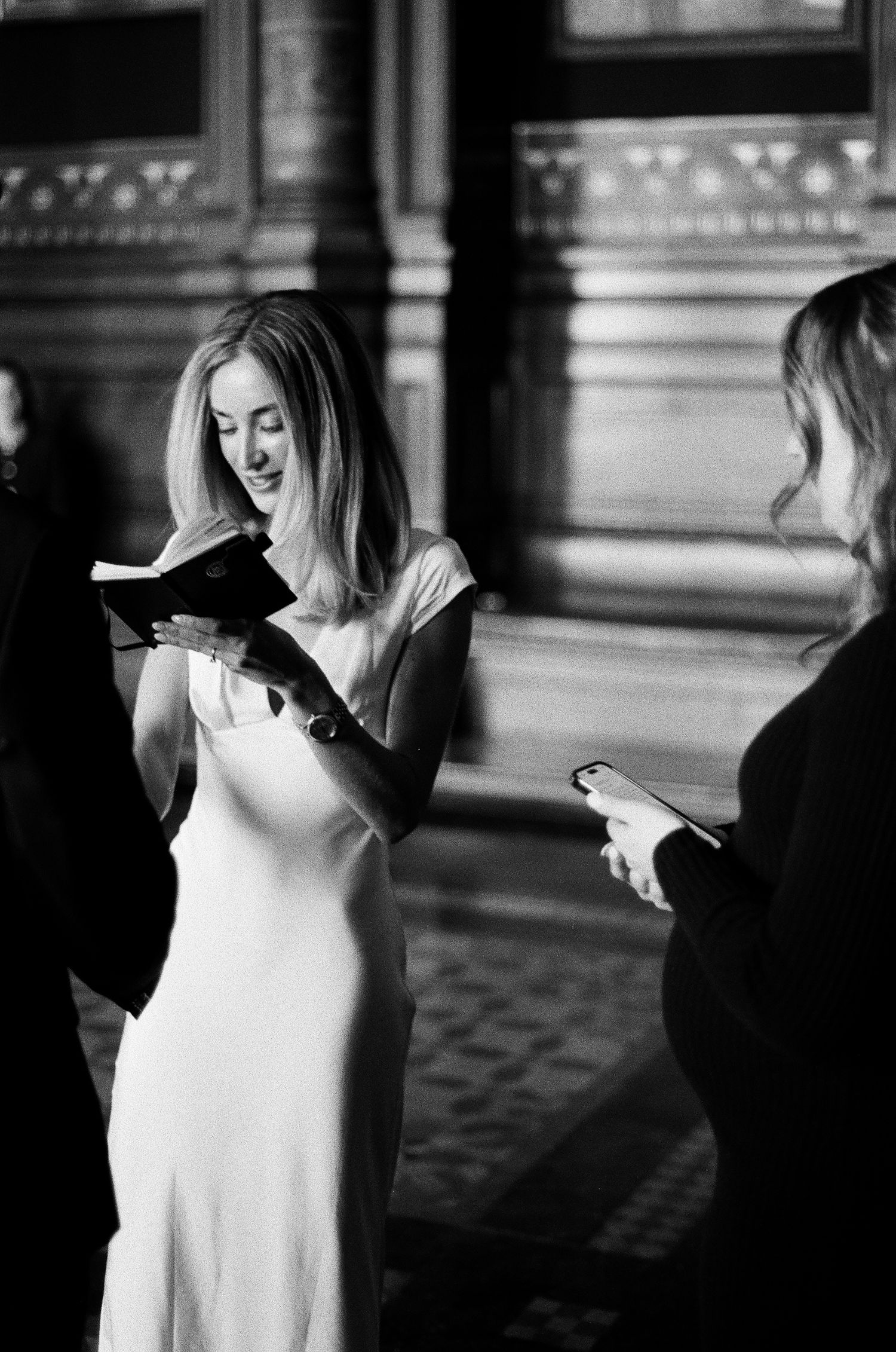 Smiling bride in white satin dress reads from book during intimate city hall wedding ceremony.Grainy film-inspired portrait