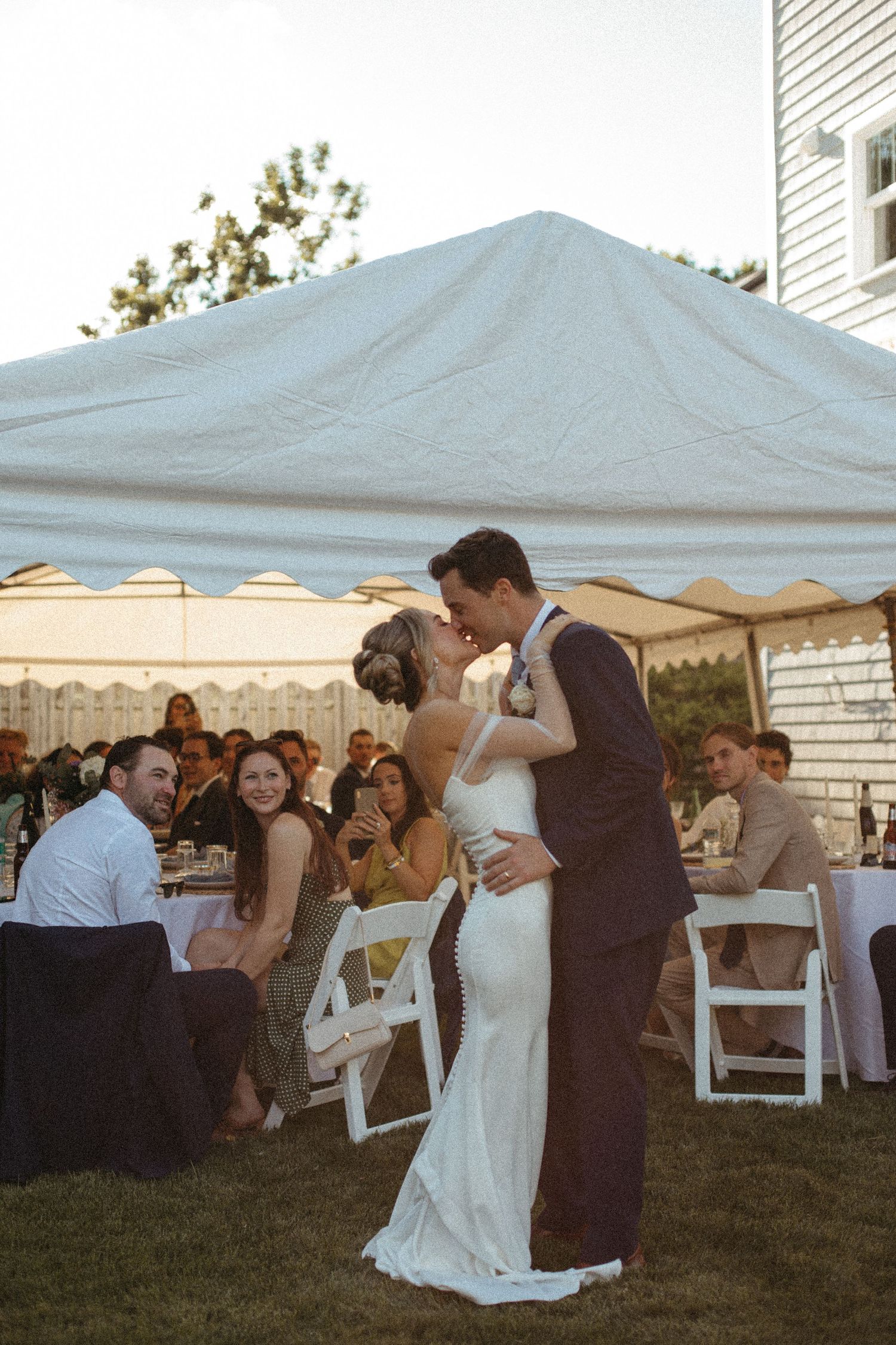 Bride and groom sharing a romantic kiss outdoors under a white tent during their wedding reception at golden hour.