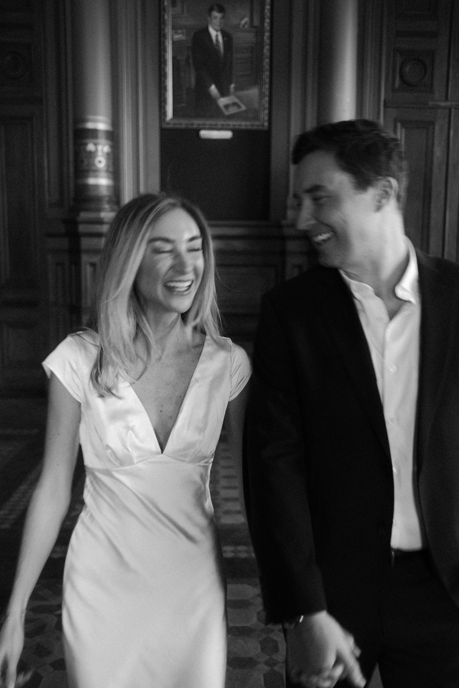 Black and white photo of a laughing bride and groom walking together in an elegant interior.