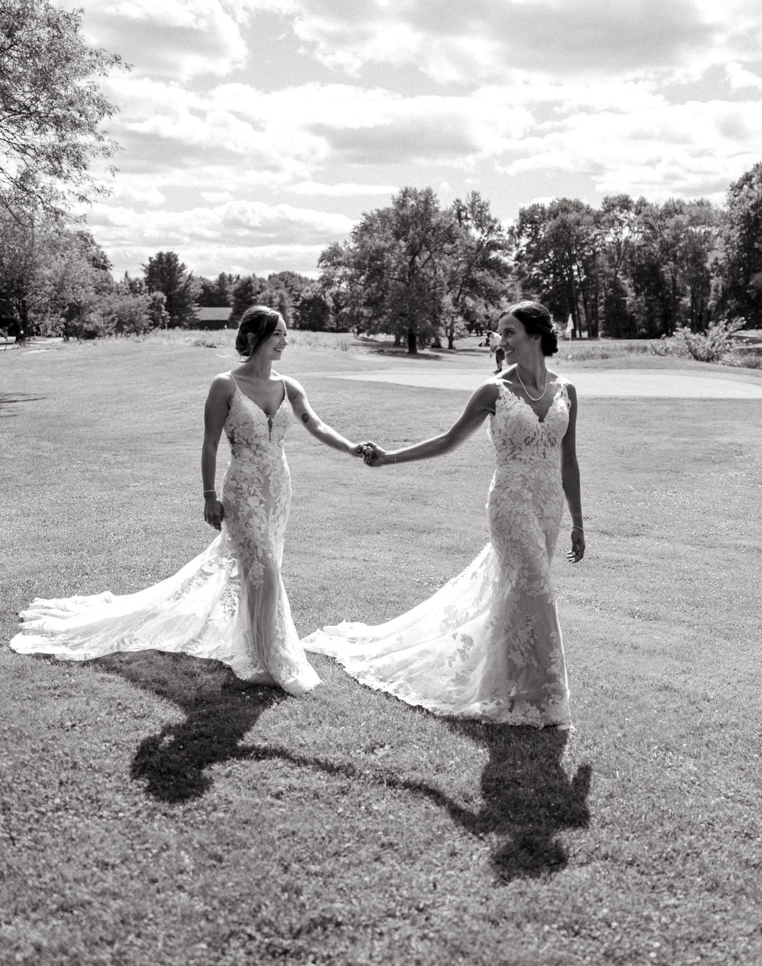 Two brides in matching lace mermaid gowns hold hands on a sunny lawn in a black and white photo.