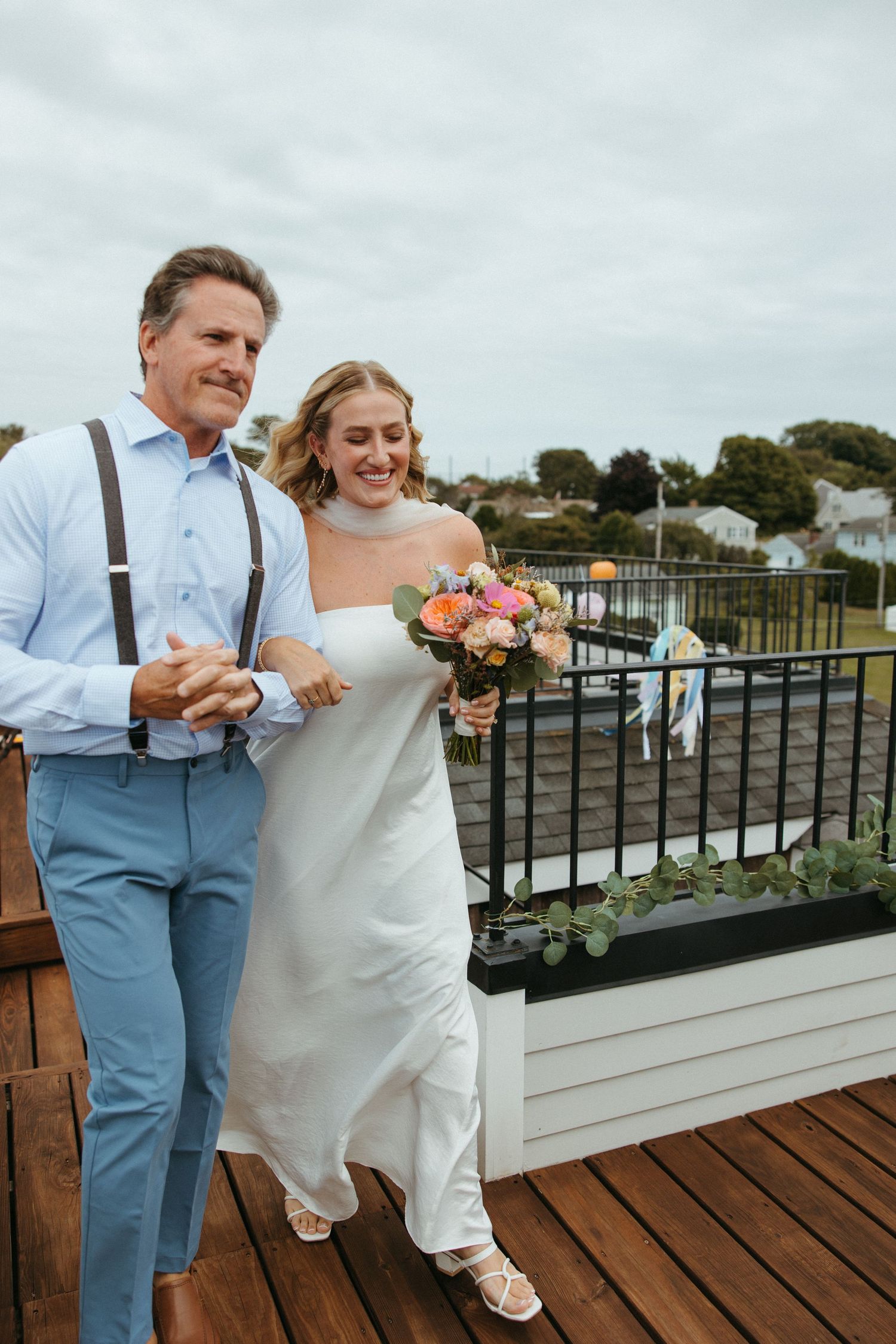 Bride holding colorful bouquet walks arm-in-arm with father in suspenders on outdoor deck.