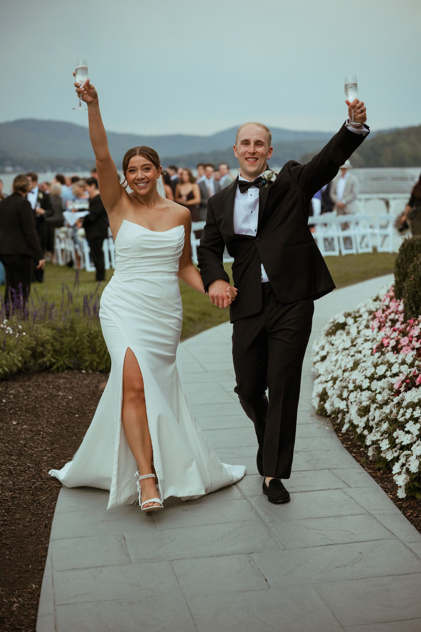 Bride and groom raise champagne glasses in celebration walking down garden path after wedding.