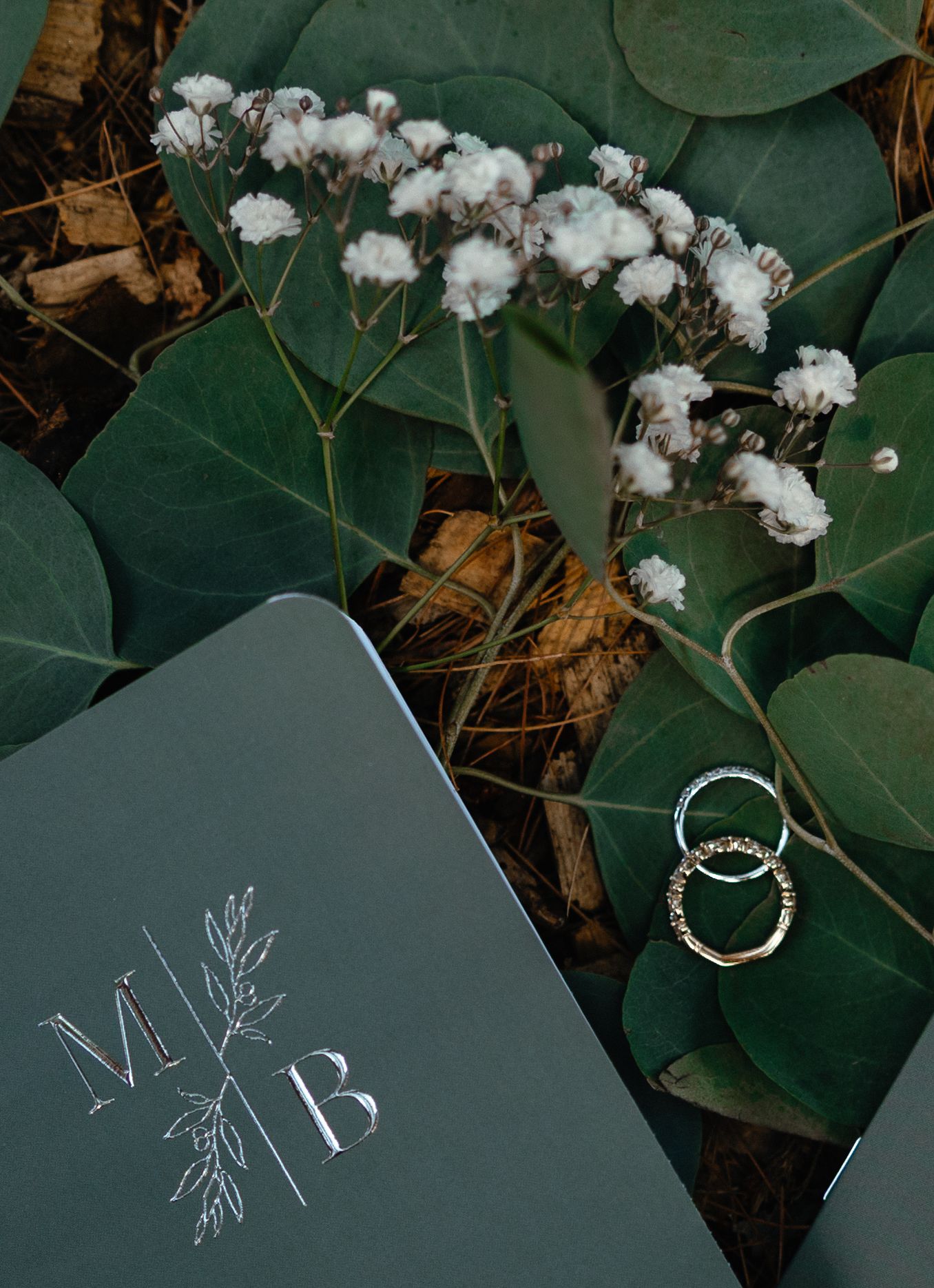 Wedding rings and monogrammed MB vow book arranged with eucalyptus and white flowers on ground.