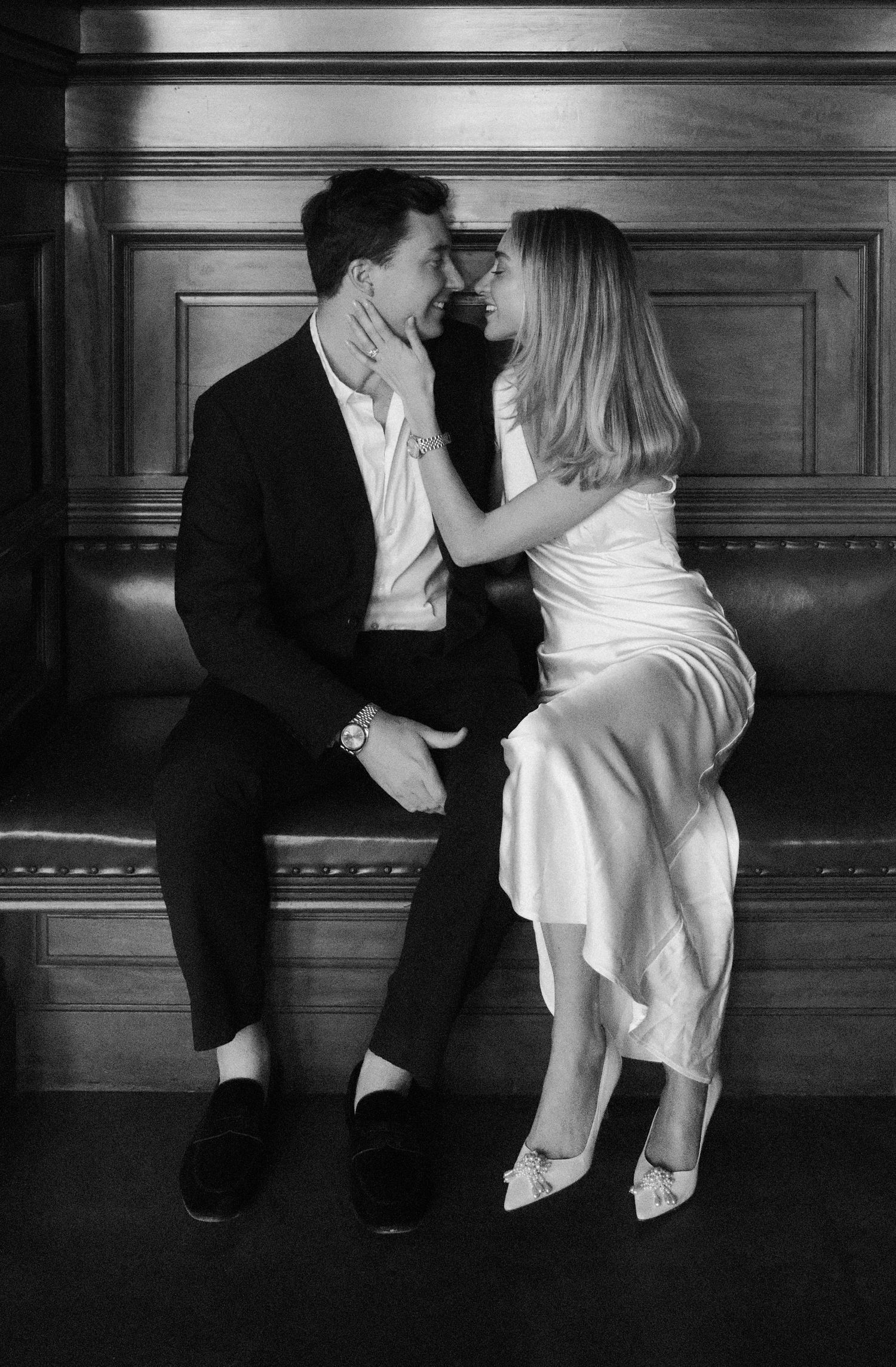 Bride in white satin dress and groom in black suit sharing a romantic moment on a bench.film-inspired