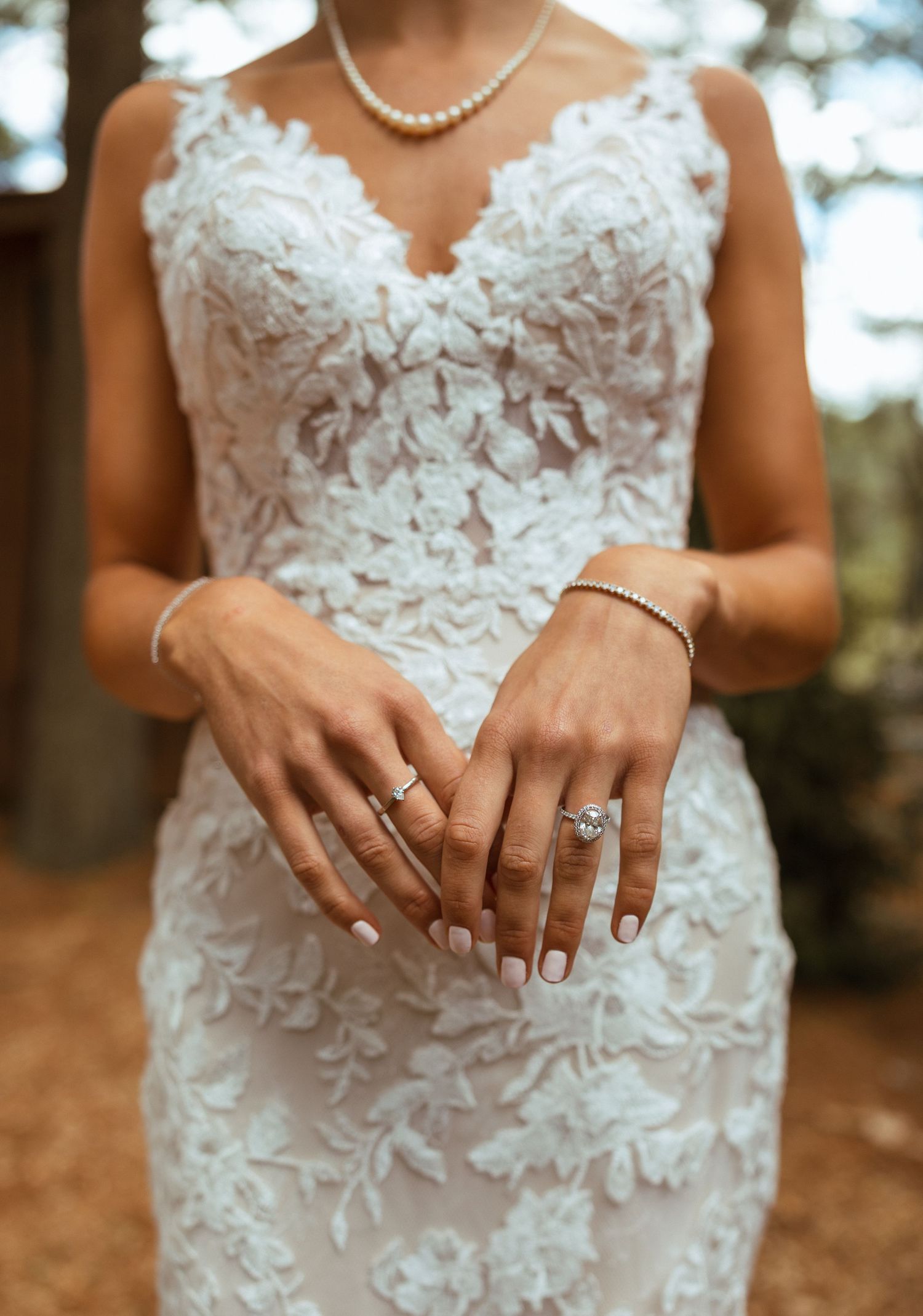 Close-up of bride in lace gown and pearl necklace displaying diamond engagement and wedding rings.