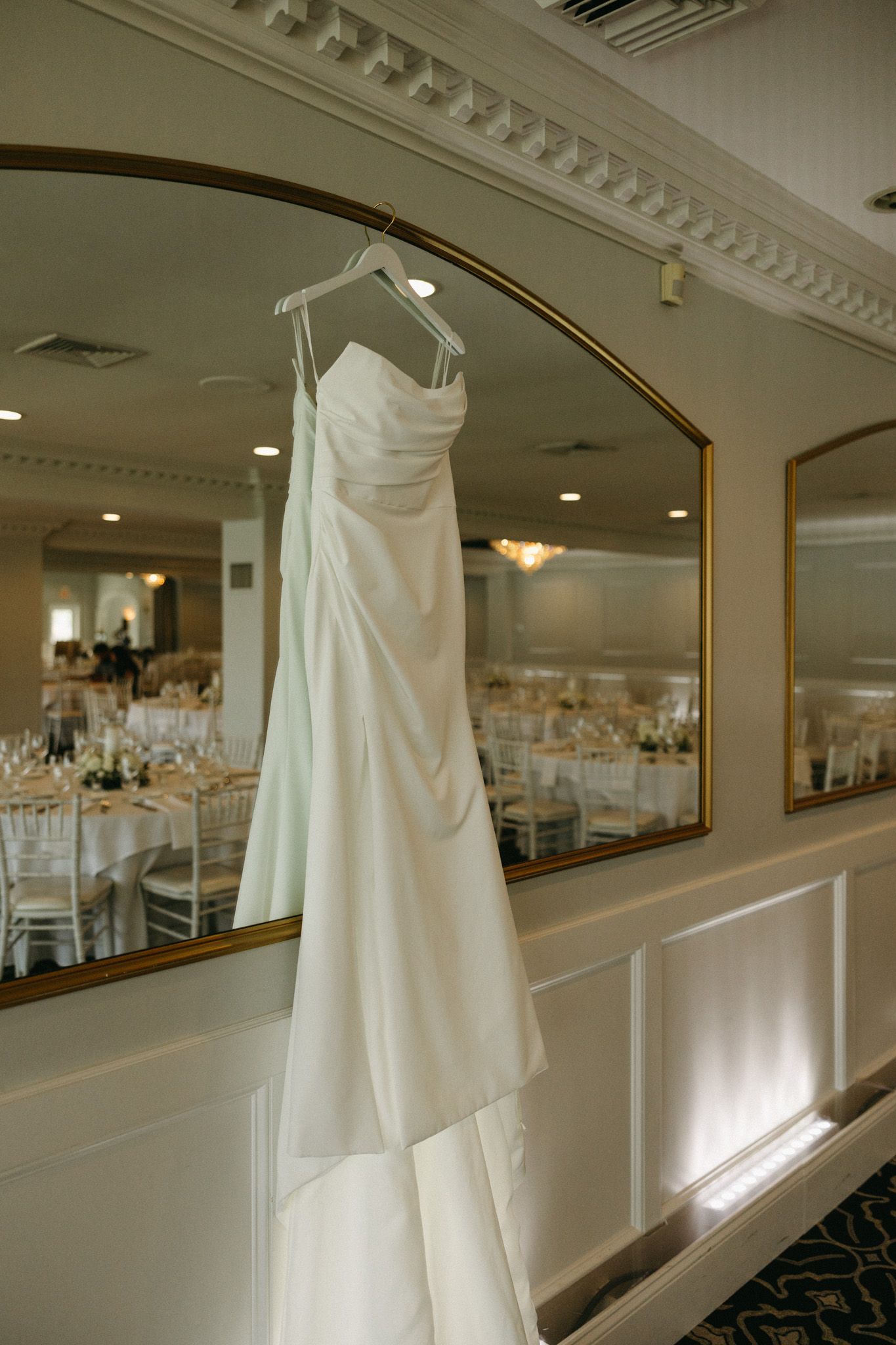 Strapless white wedding gown hanging on hanger in front of gold-framed mirror in venue.