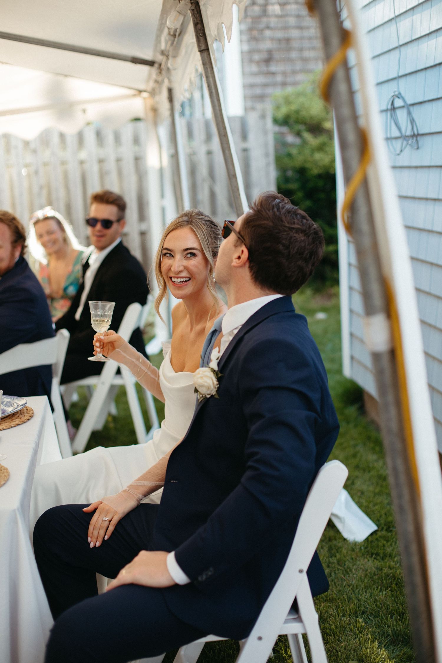 Radiant bride raising champagne glass and laughing with groom at outdoor wedding reception.