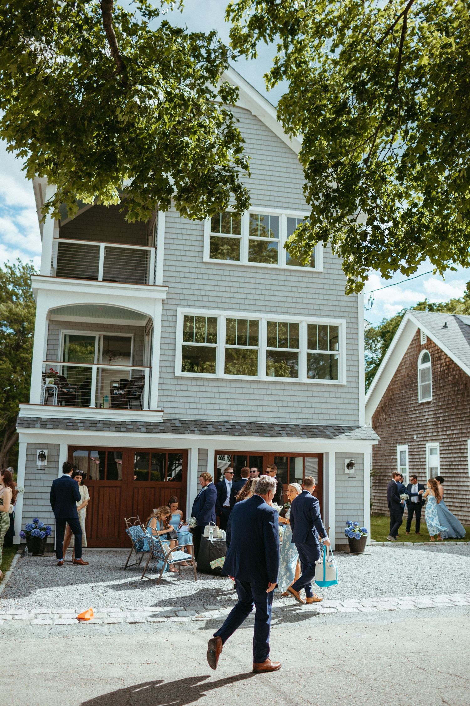 Wedding guests gather outside a three-story gray shingle coastal home on a sunny day.