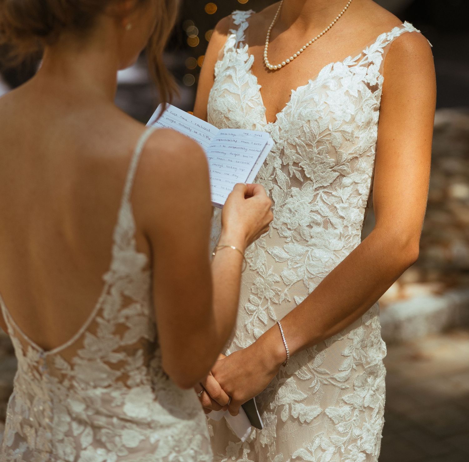 Two brides in lace wedding gowns exchange handwritten vows during outdoor wedding ceremony.