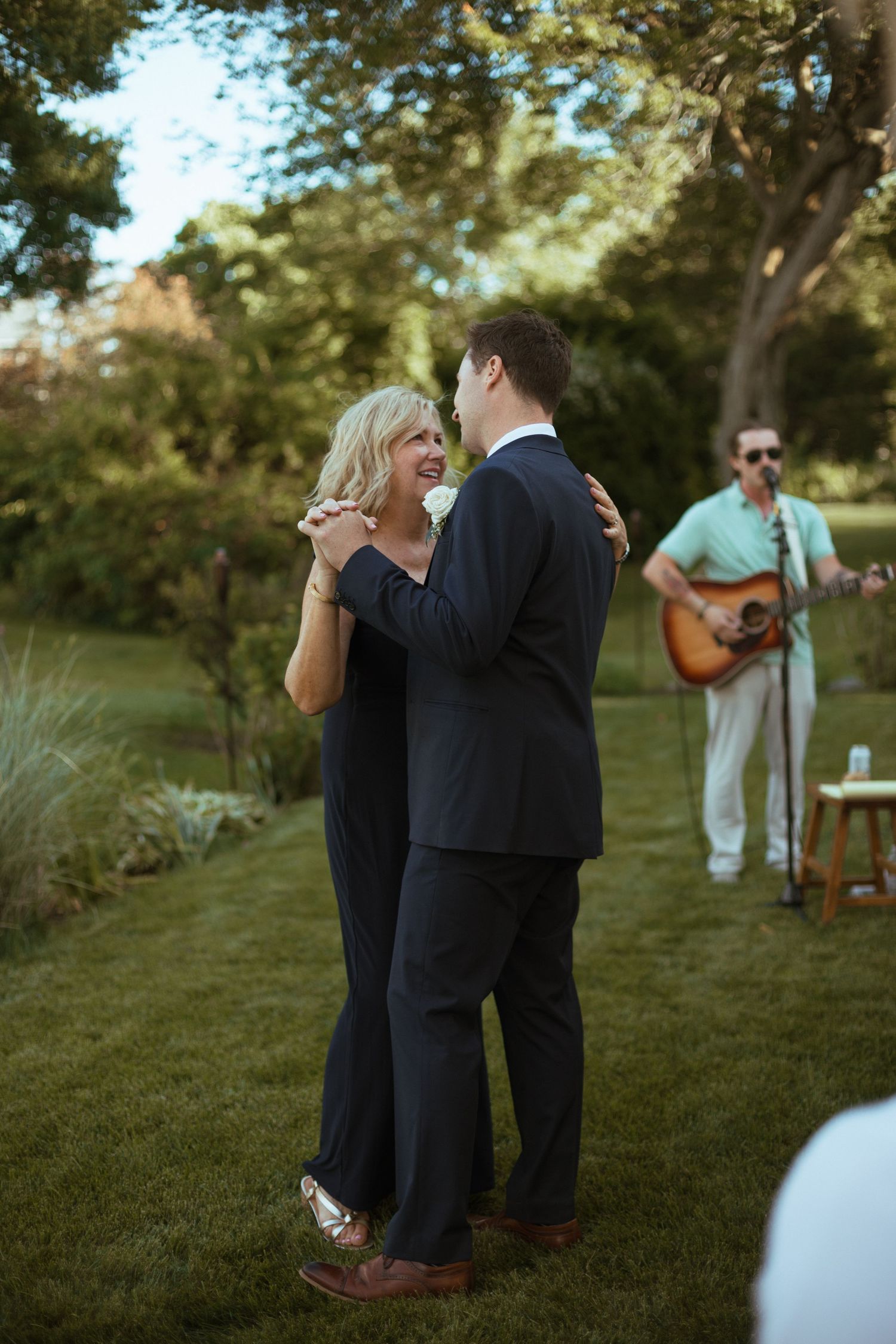 Groom dancing with mother in navy dress at outdoor wedding with live guitarist performing.