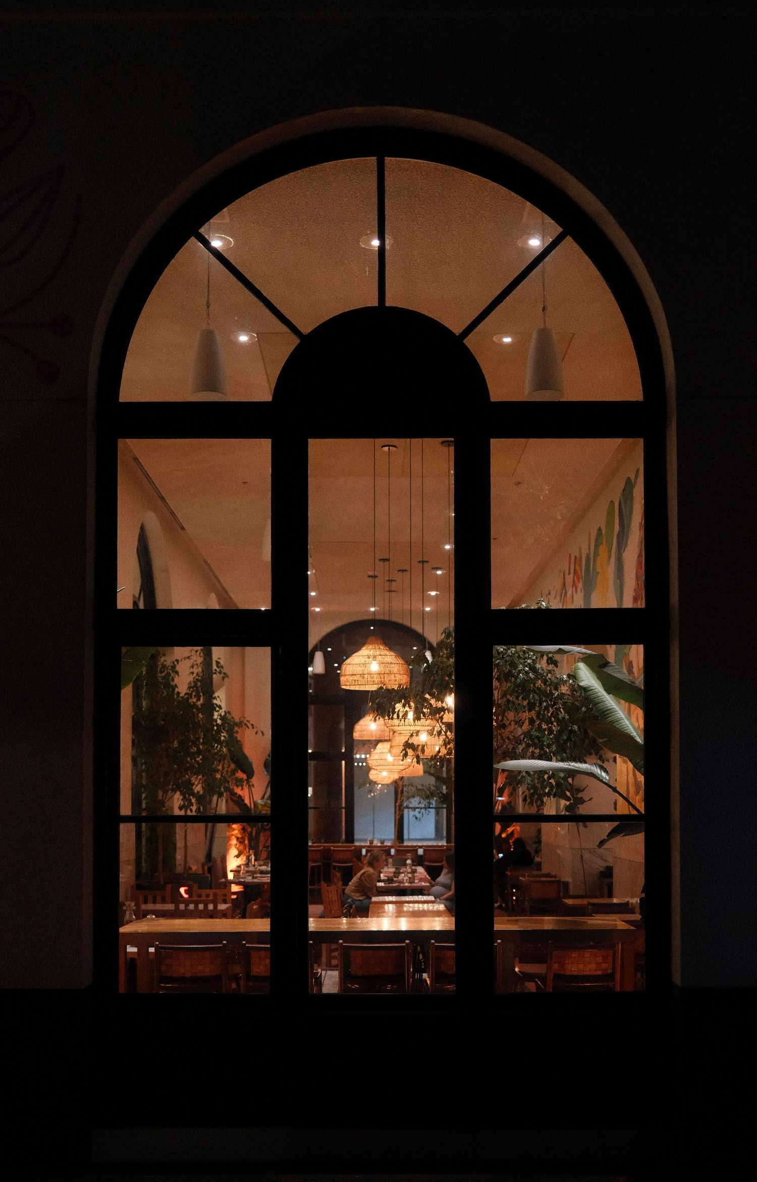 Elegant arched window at night reveals warmly lit restaurant interior with hanging lights and lush plants.