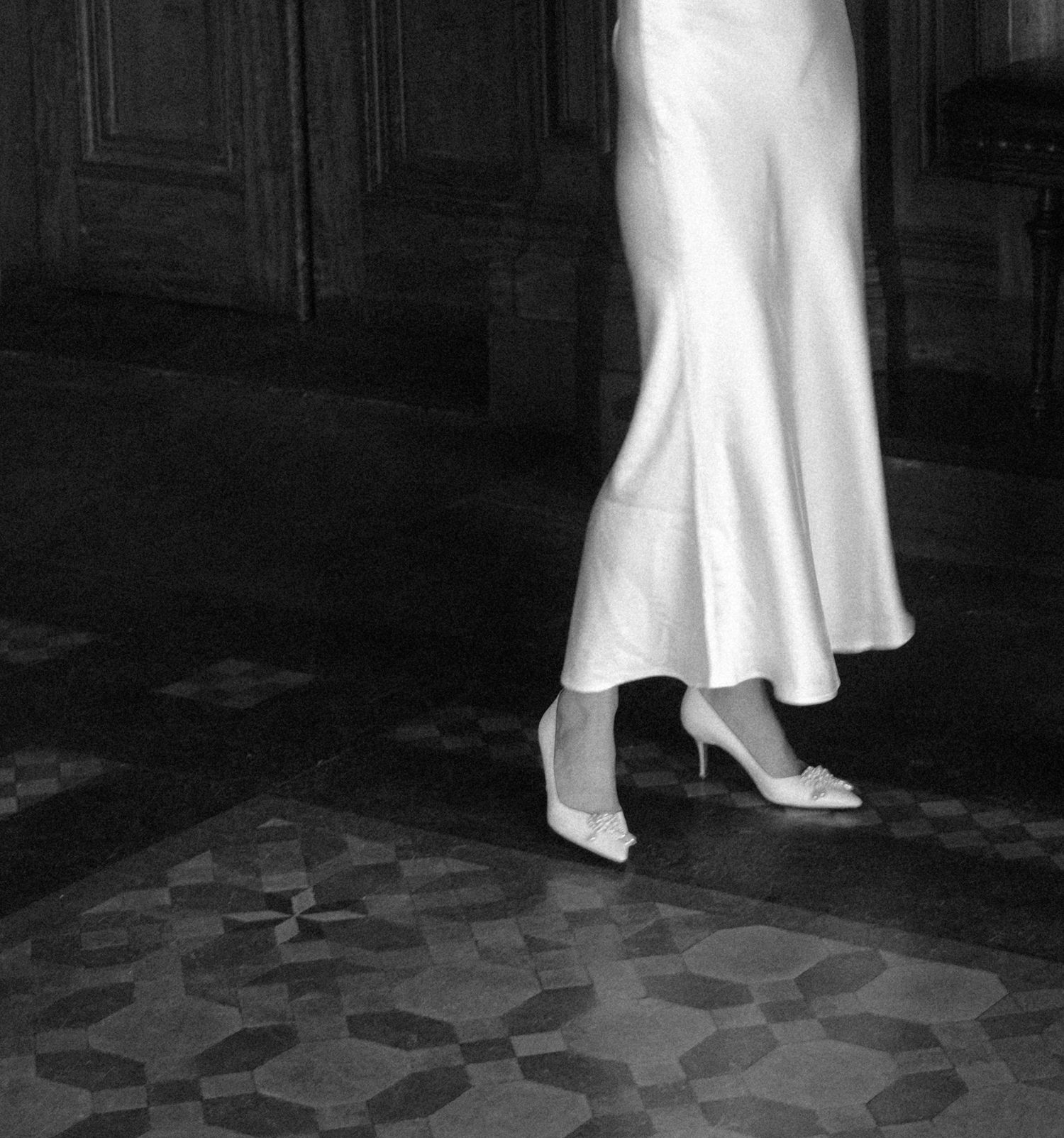 Close-up of bride's white satin dress and heels on ornate tiled floor.film-inspired