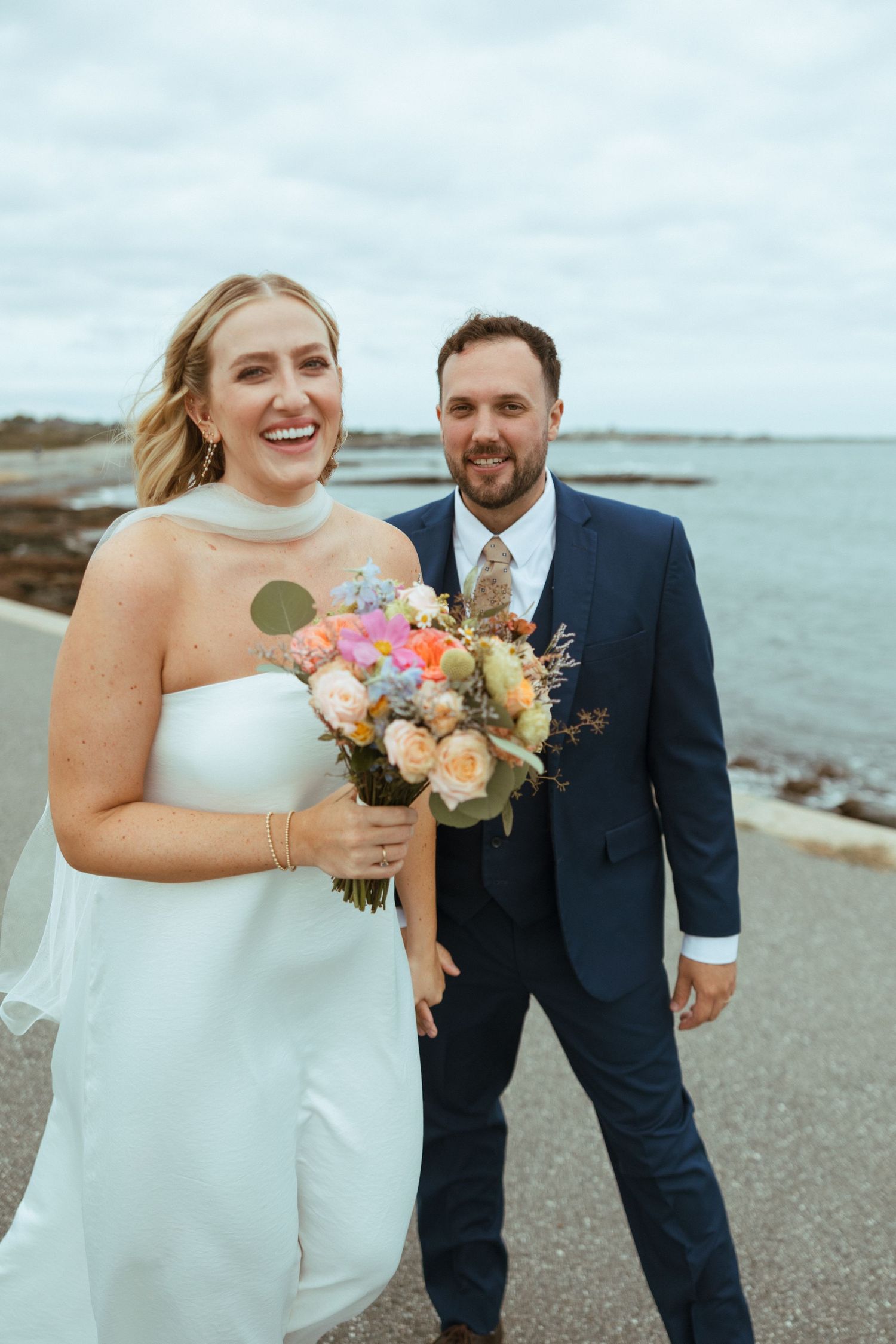 Laughing bride in white gown with veil holds vibrant bouquet beside smiling groom in navy suit along a coastal path.