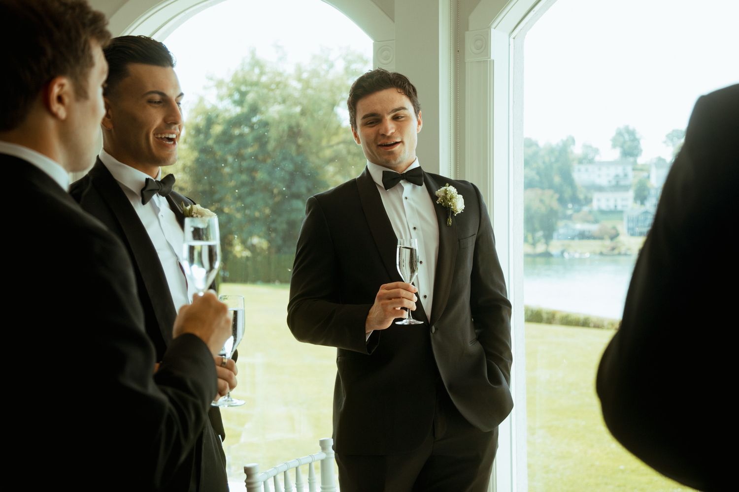 Groomsmen in black tuxedos holding champagne glasses chatting near window at wedding.