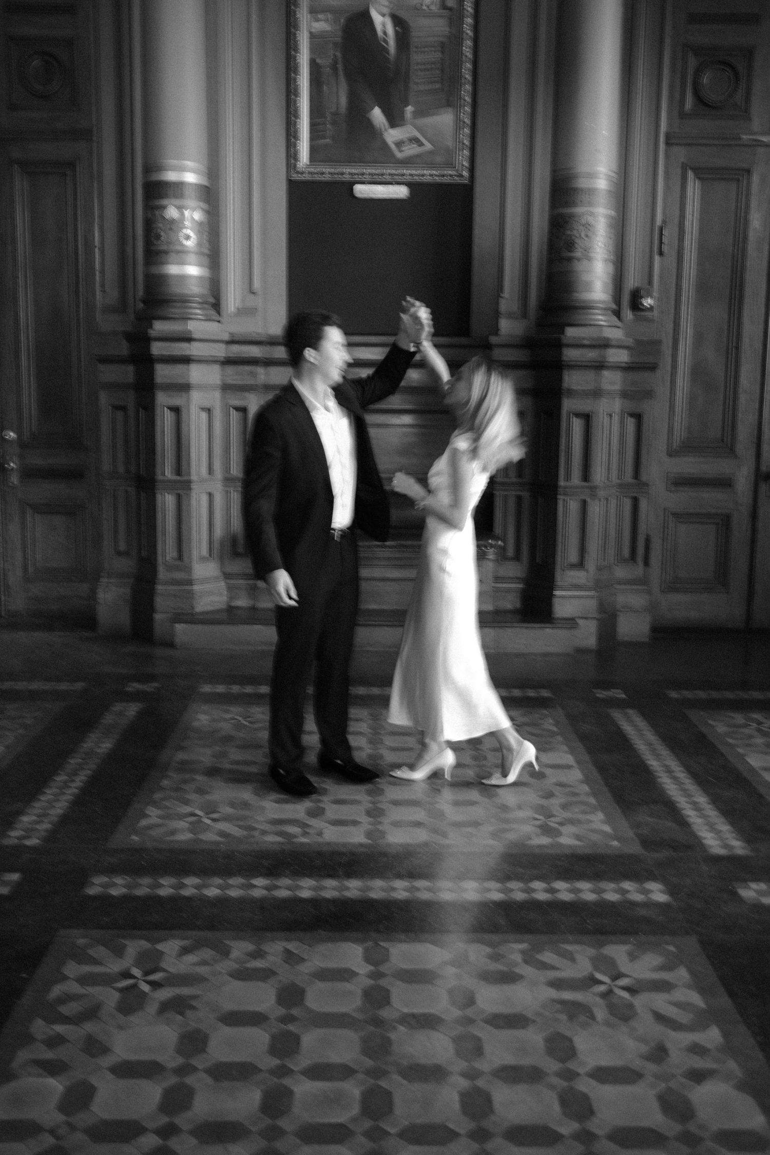 Black and white photo of a couple dancing elegantly in an ornate hall, man in suit twirling woman in white dress.film-inspired