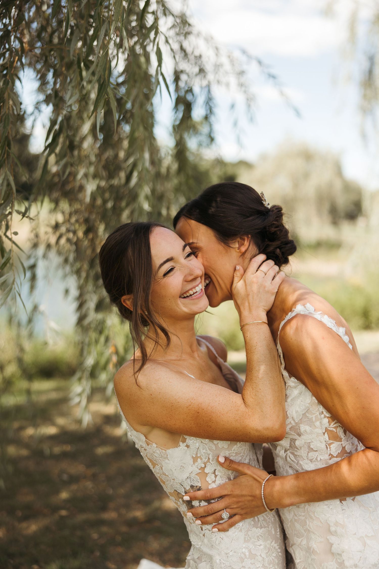 Two brides laugh joyfully as one whispers in the others ear under willow tree branches outdoors.