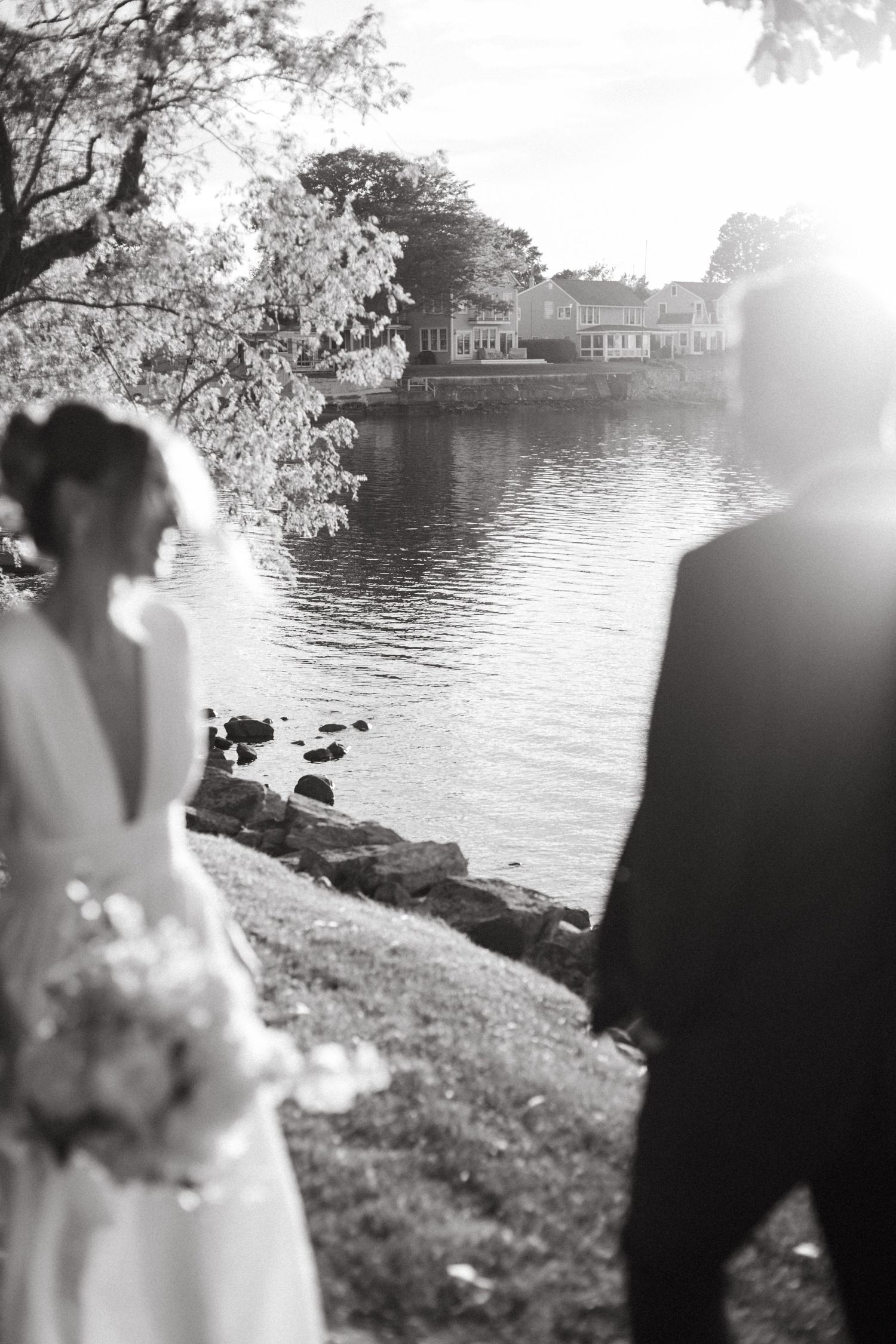 Black and white photo of bride and groom holding hands by sunlit waterfront.