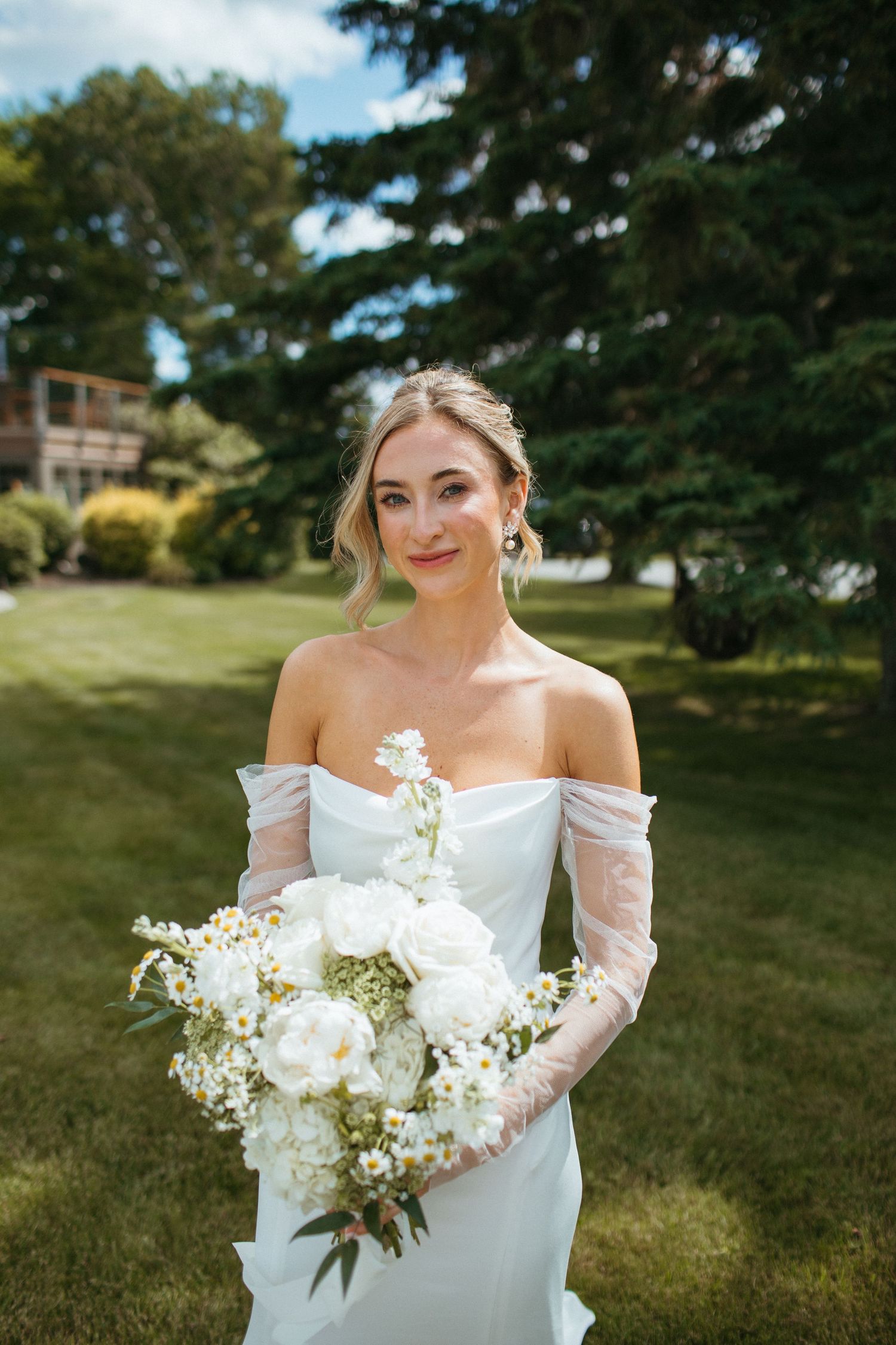 Bride in white off-shoulder gown with sheer gloves holding large white peony and baby's breath bouquet.