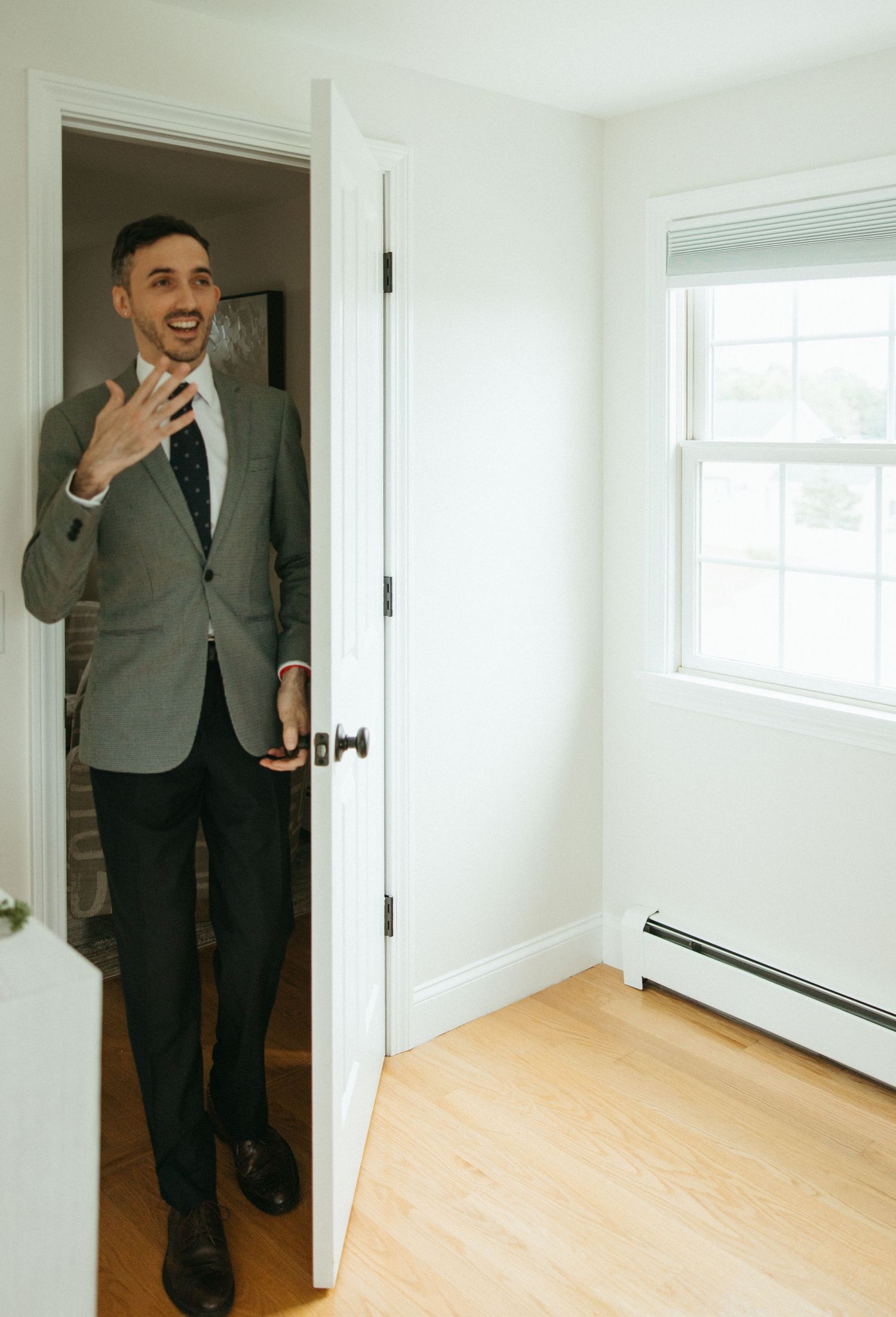 Smiling groom in grey suit peeking through doorway during wedding day preparations.
