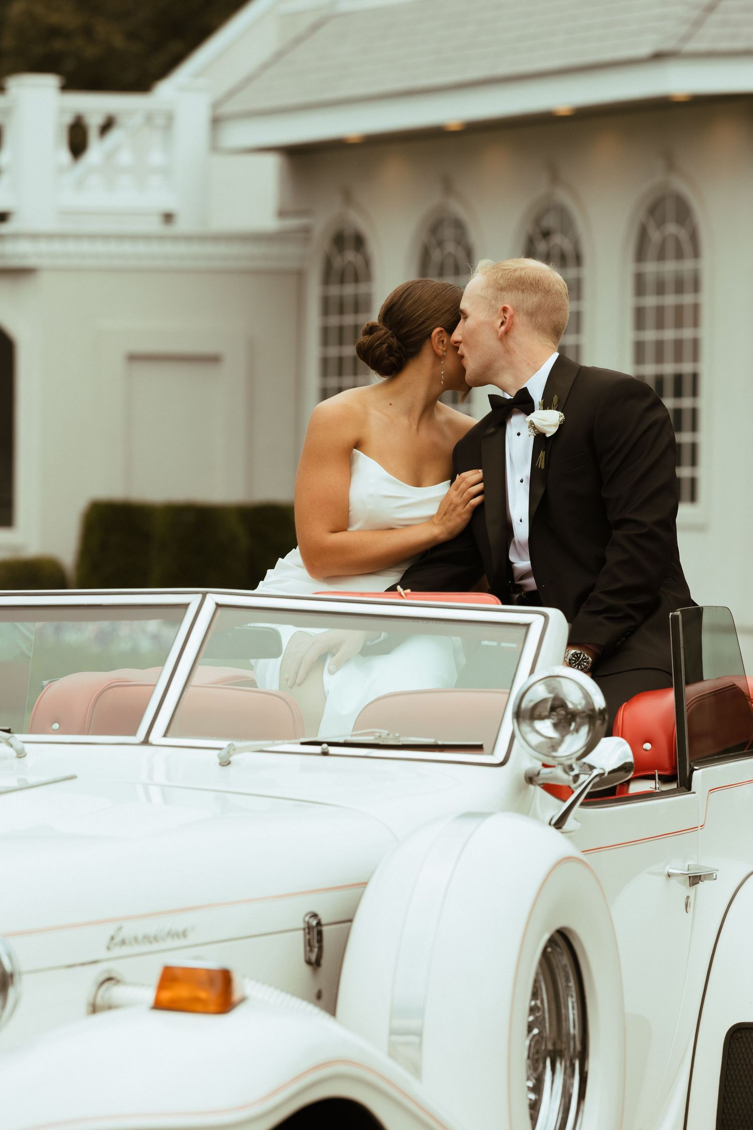 Bride and groom sharing a kiss while seated in a classic white Excalibur convertible wedding car.