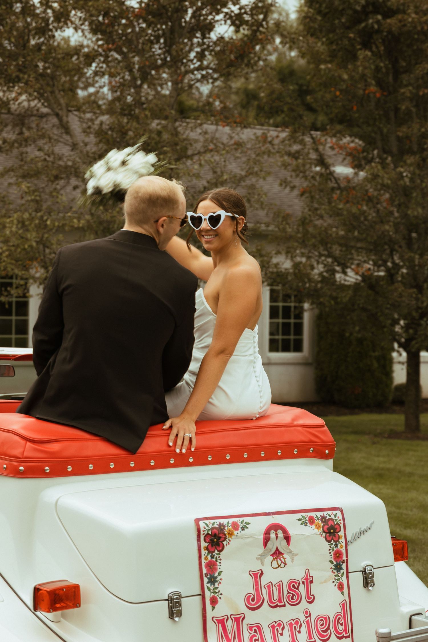 Bride in white dress with heart sunglasses and groom sit atop a Just Married vintage convertible car.