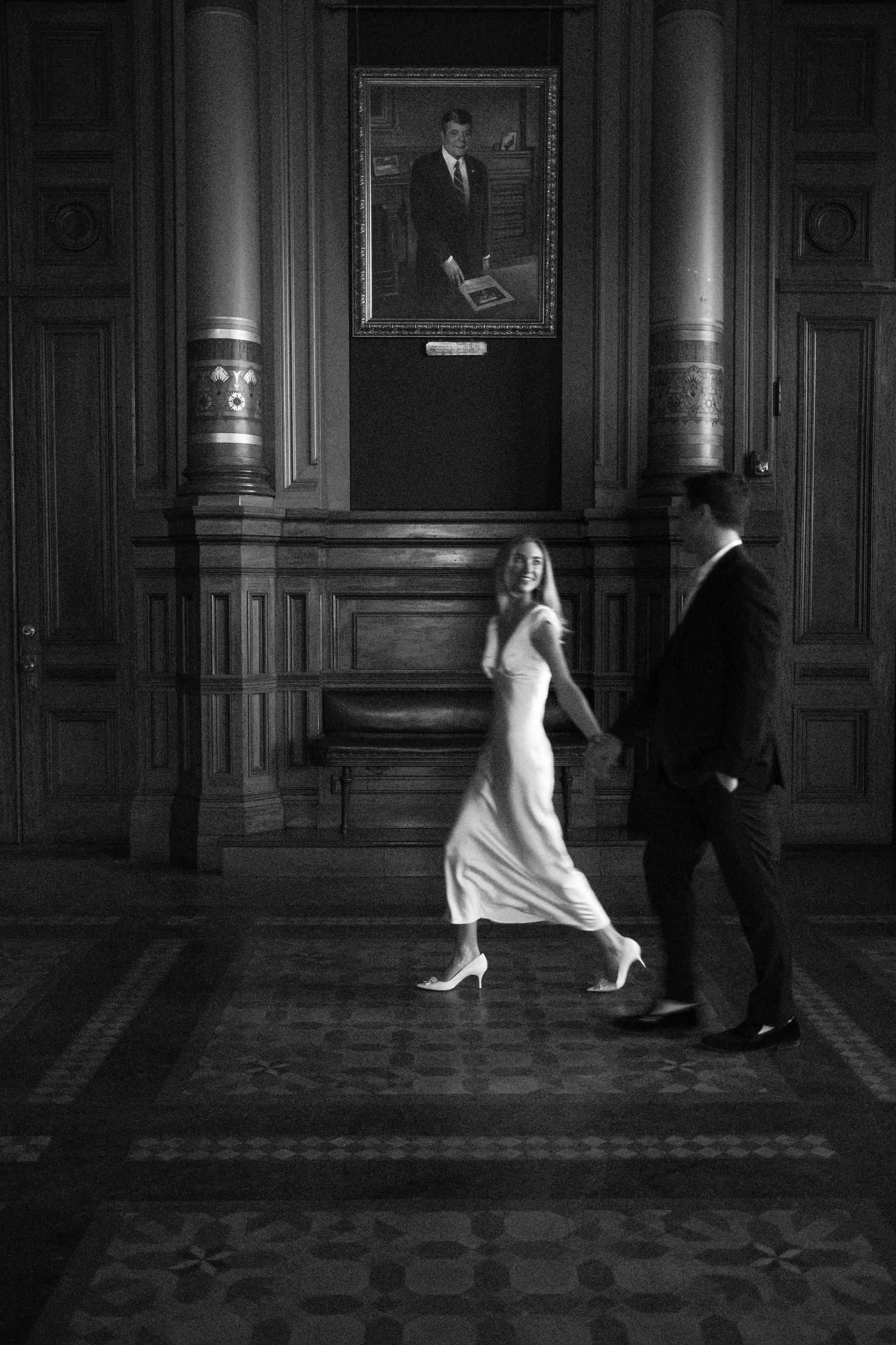 Black and white photo of a couple walking hand in hand through an elegant wood-paneled hall with portrait above.film-inspired