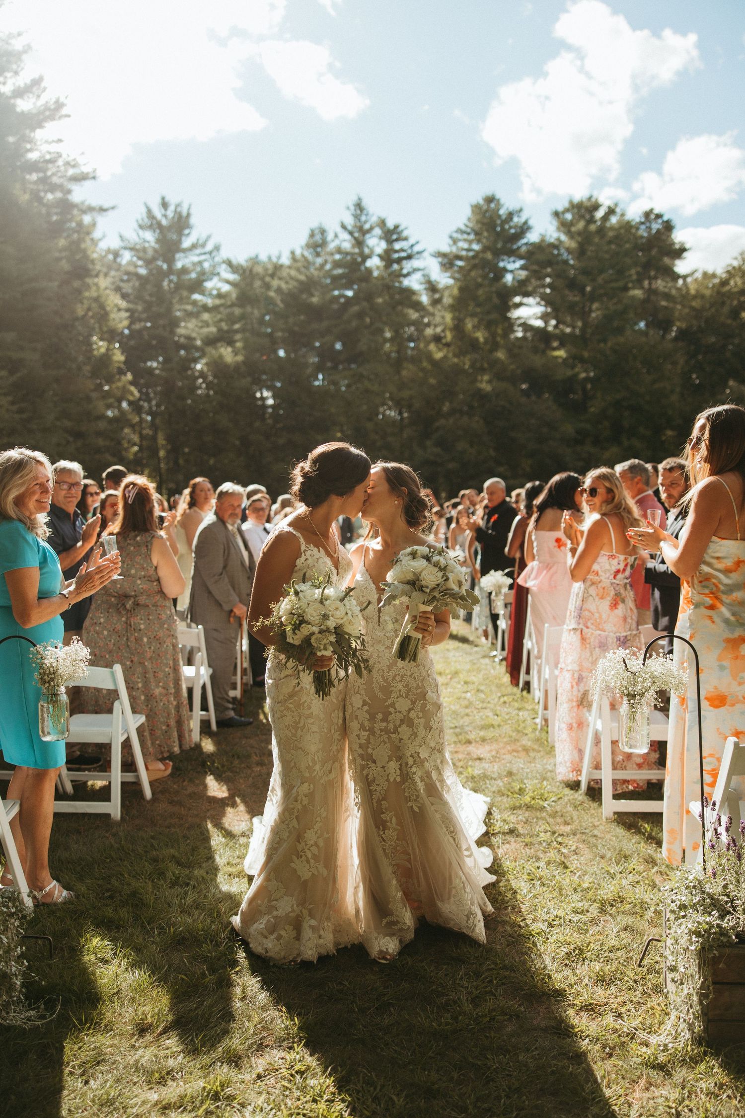 Two brides share first kiss walking back down aisle as guests celebrate at outdoor wedding.