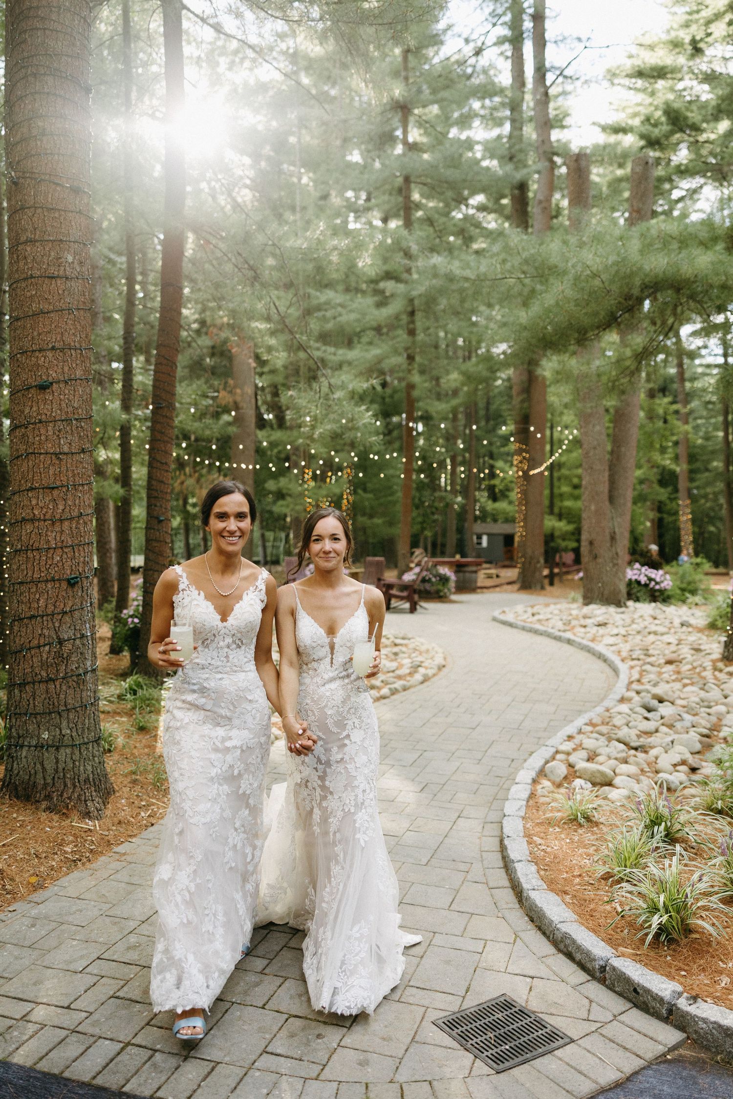 Two smiling brides in lace wedding gowns walk hand in hand along a forest path with string lights.
