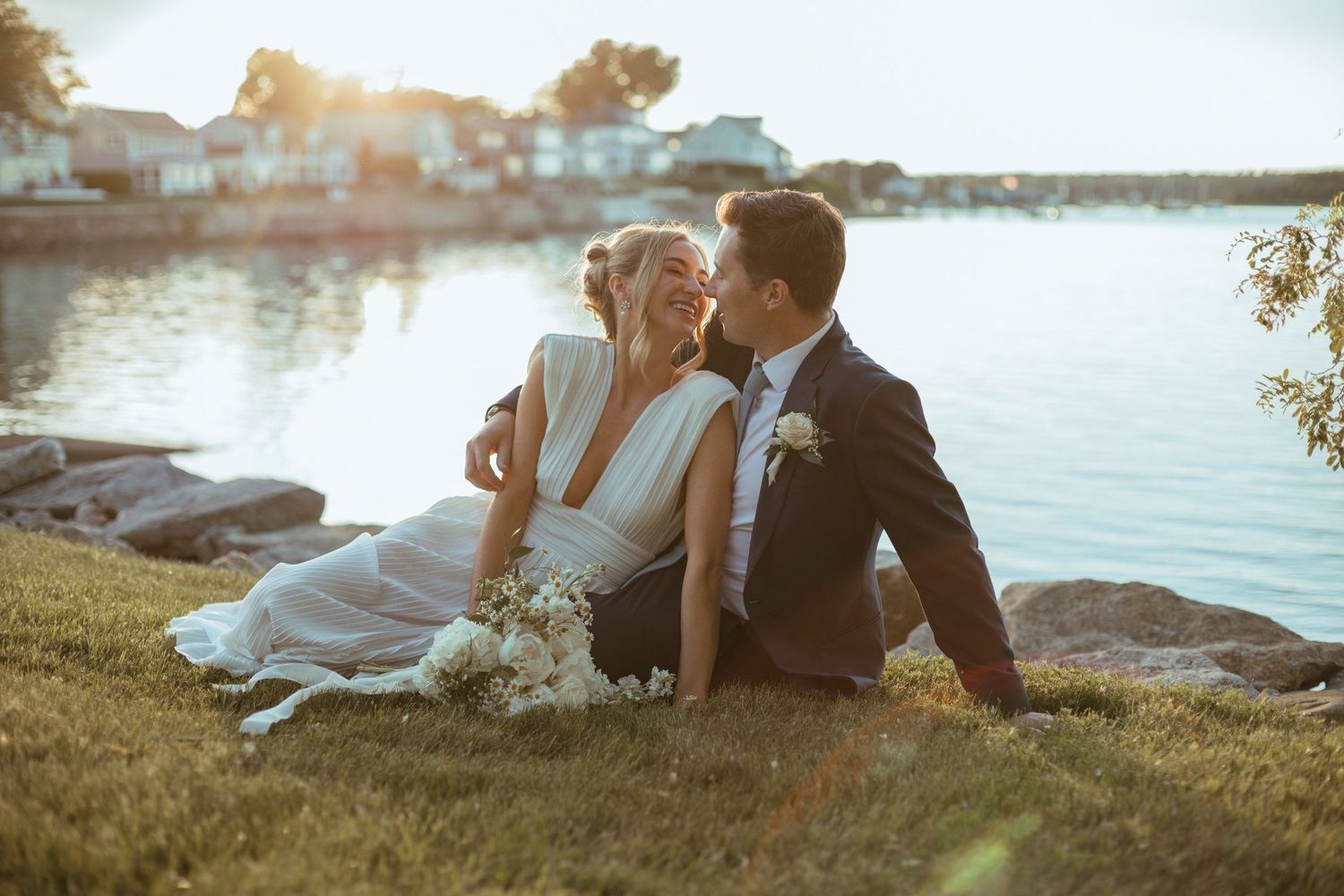 Bride and groom sitting by a sunlit waterfront, sharing a romantic moment on their wedding day.