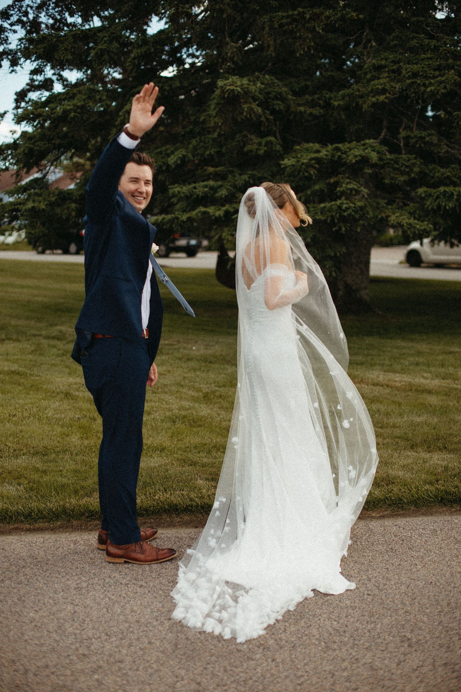 Groom waving while bride's veil flows in wind during outdoor wedding portraits.
