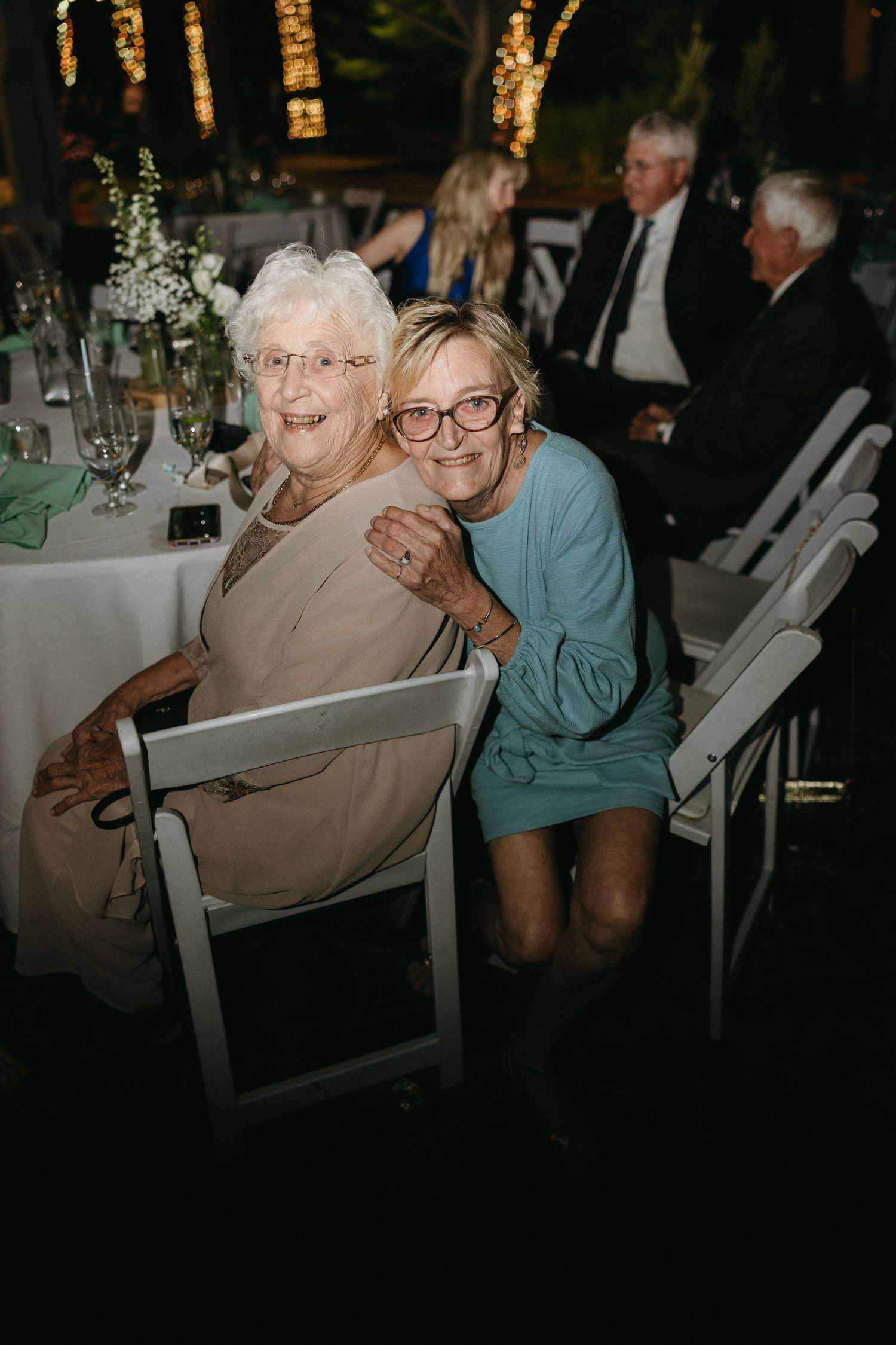 Two women smile together at wedding reception seated at decorated table.film-inspired
