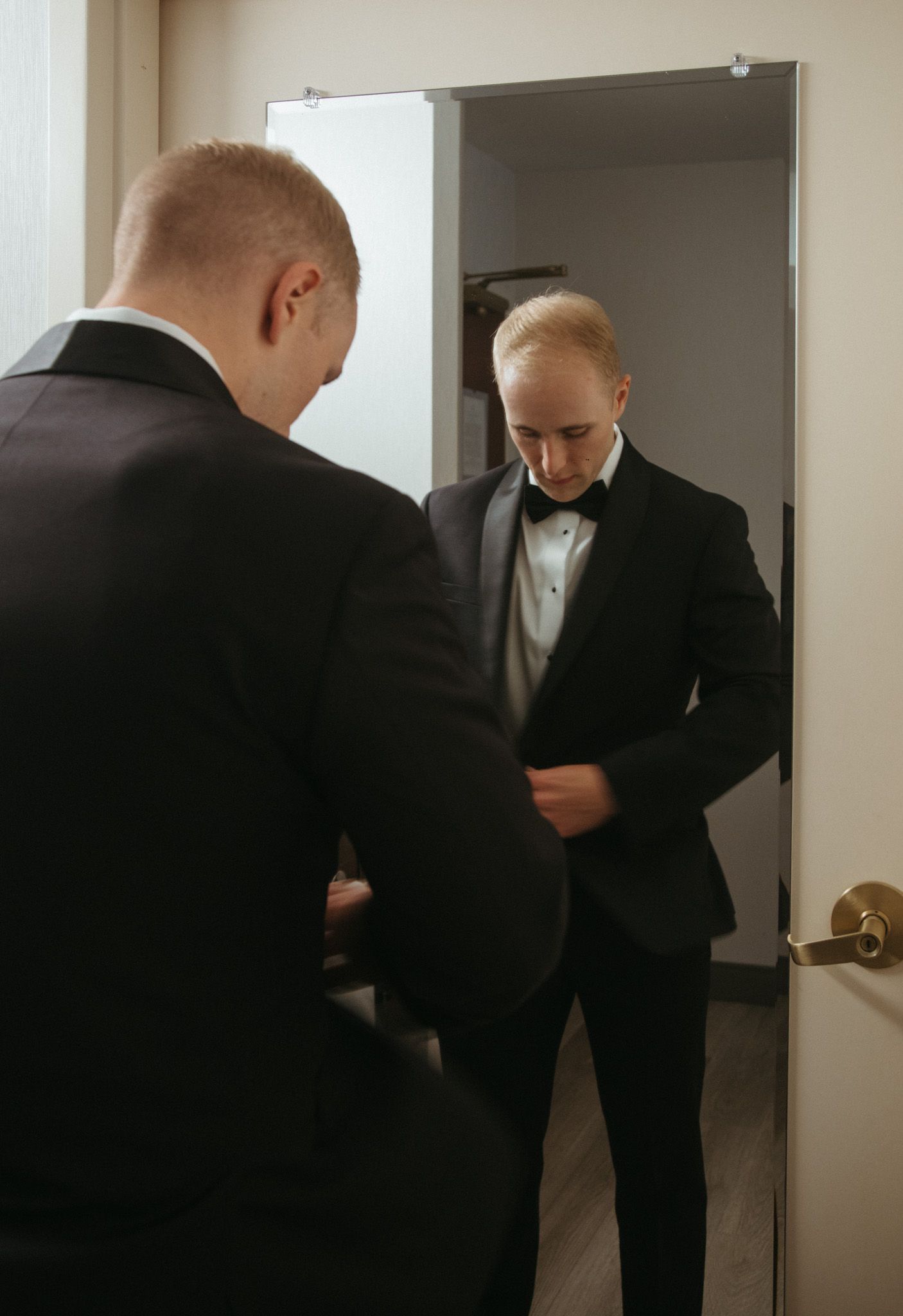 Groom in black tuxedo buttoning jacket while looking in mirror before wedding ceremony.