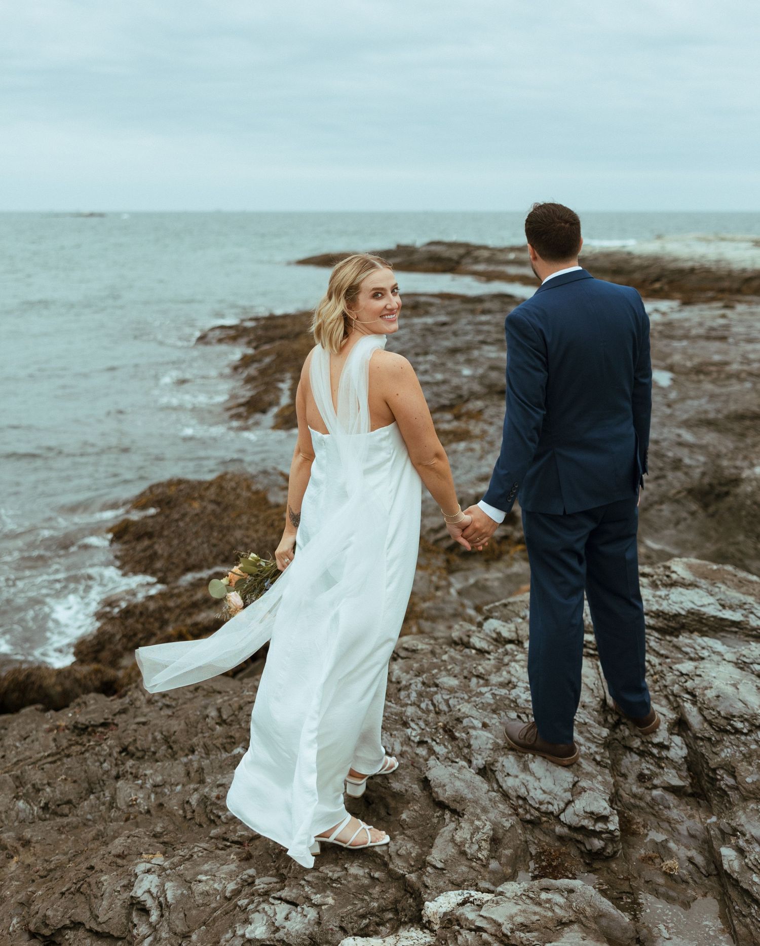 Bride in white halter gown looks back smiling while holding groom's hand on rocky coastal terrain under cloudy skies.