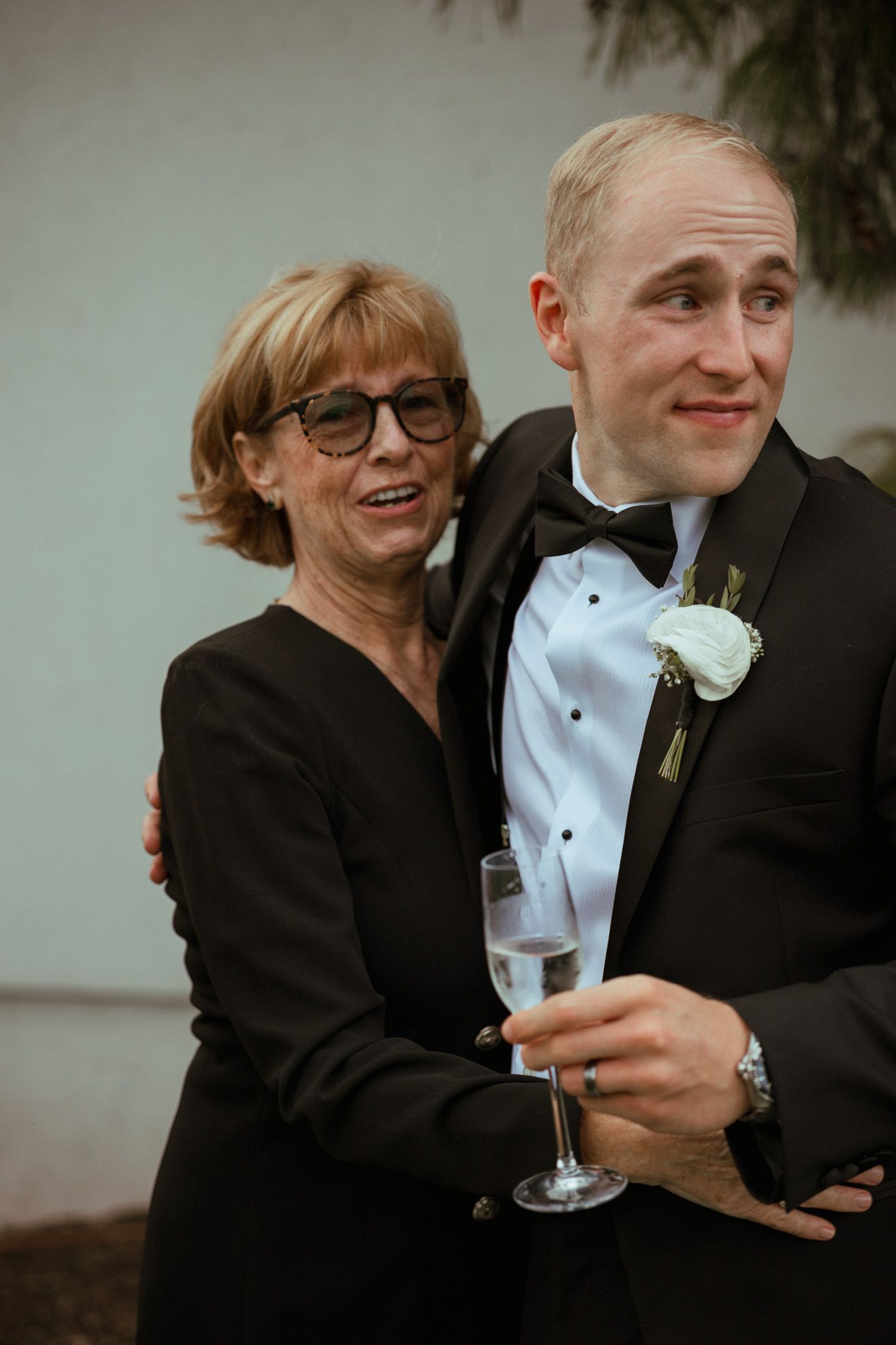 Groom in tuxedo stands with older woman who embraces him proudly at wedding reception.