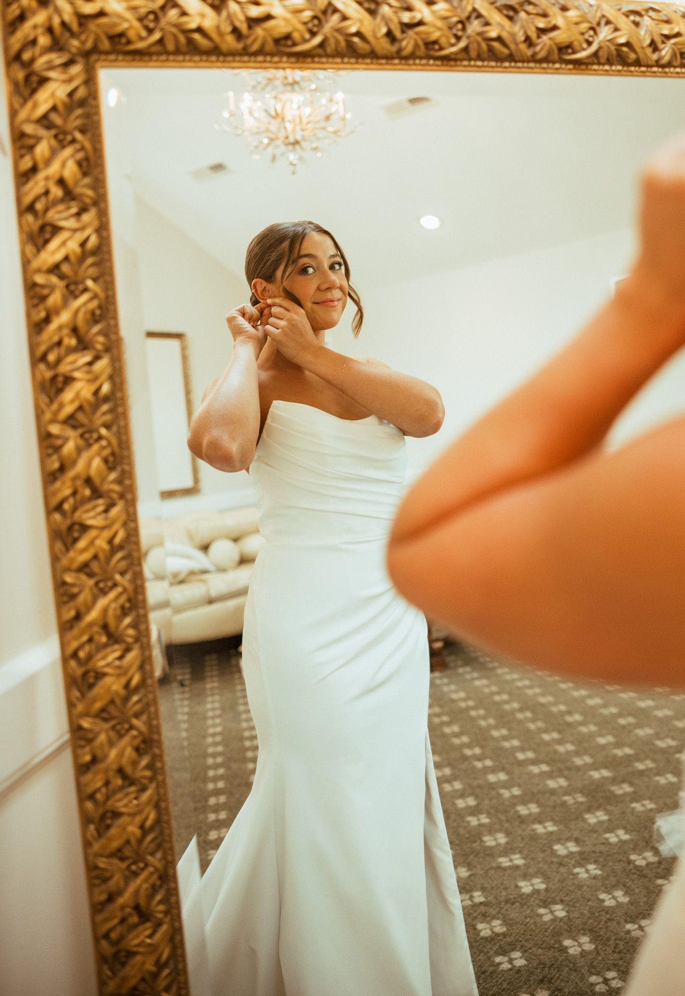 Bride in strapless white wedding gown adjusting earring in ornate gold-framed mirror.