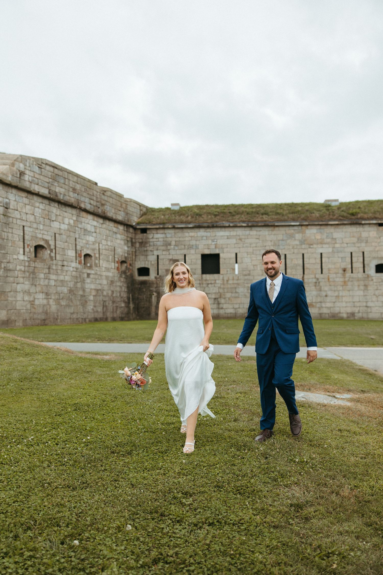 Smiling bride in white strapless gown and groom in navy suit walk hand-in-hand across green grass near a stone fort.