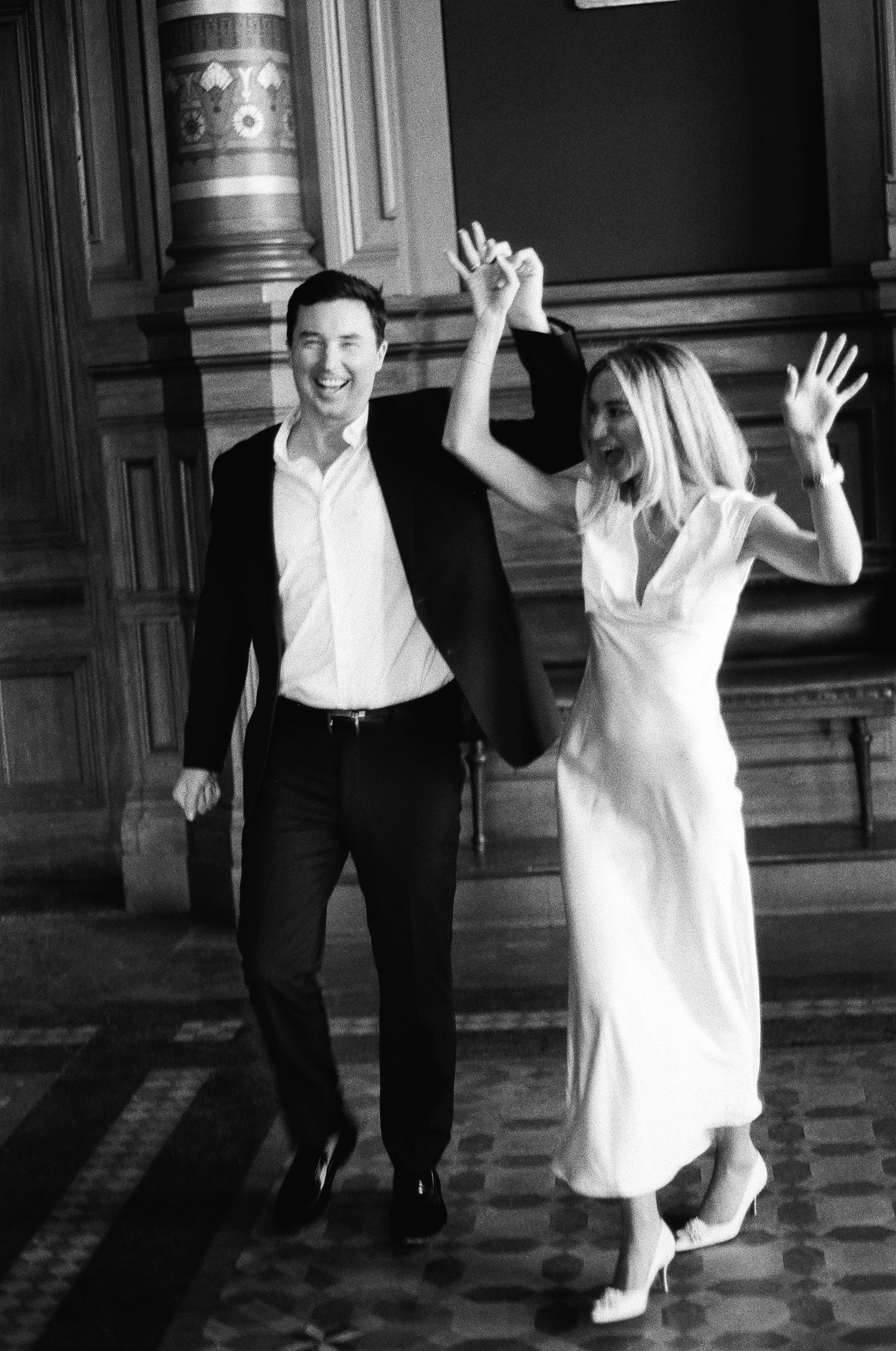 Joyful newlyweds celebrate with raised hands walking through grand city hall after wedding ceremony.