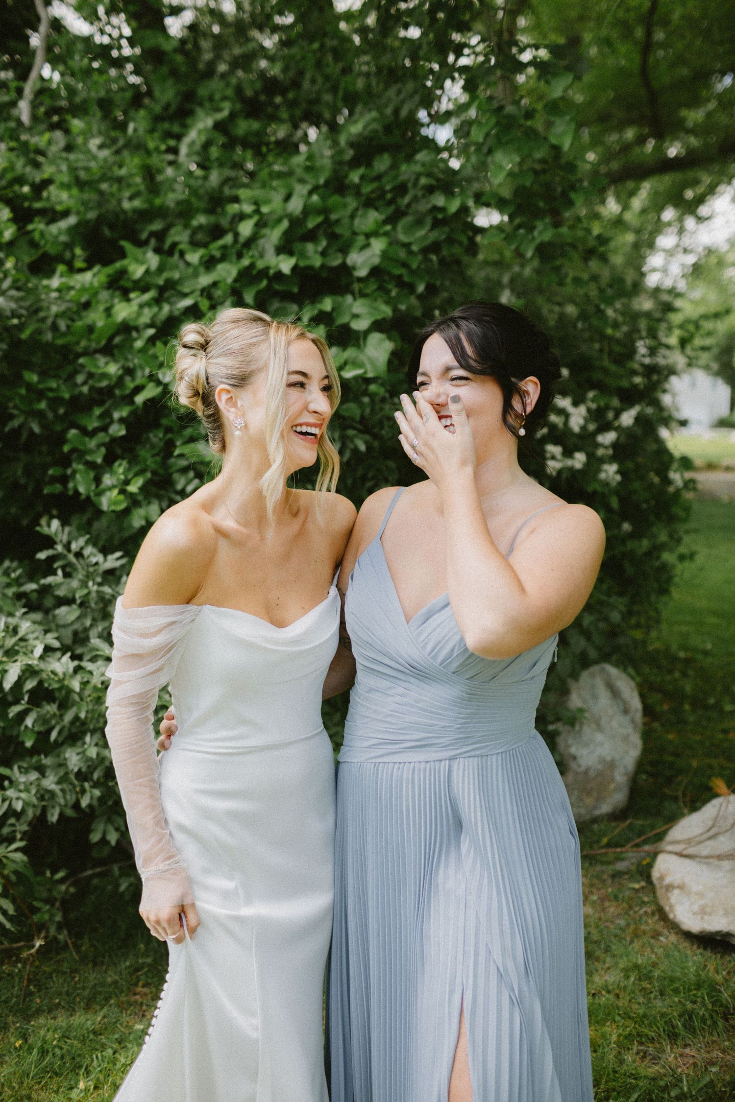 Bride and bridesmaid in blue dress laughing together in garden setting.