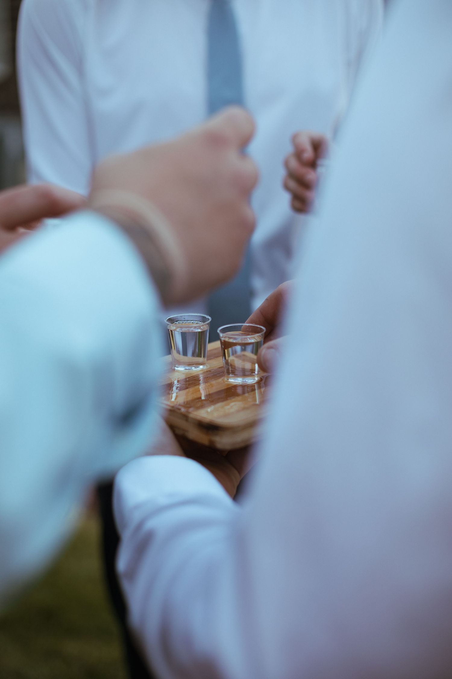 Guests holding shot glasses on wooden tray at outdoor evening wedding celebration.