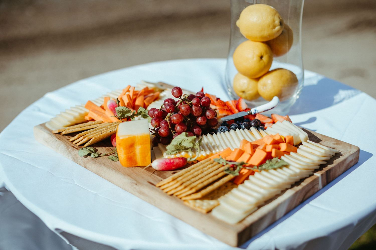 Rustic wooden charcuterie board with grapes, crackers, cheese, and fruit at outdoor wedding reception.